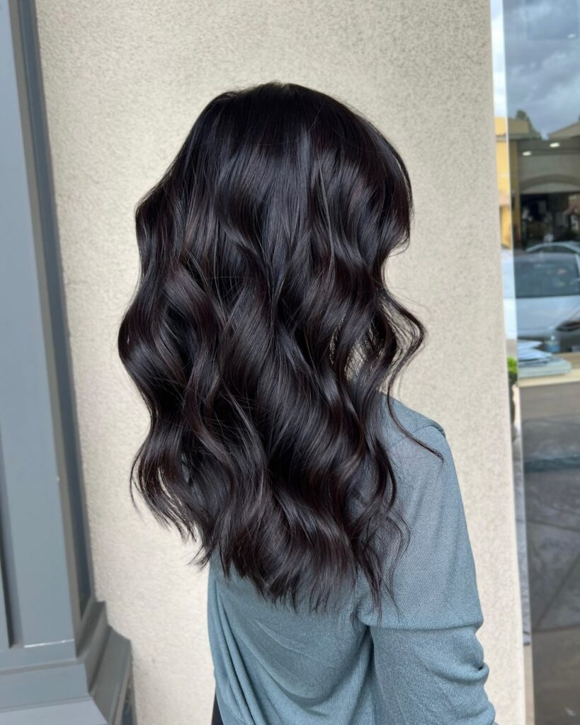Back view of a girl with long, deep espresso brown hair styled in soft waves, wearing a grey off-shoulder knit top, standing in front of a modern building with glass and concrete features on an overcast day.