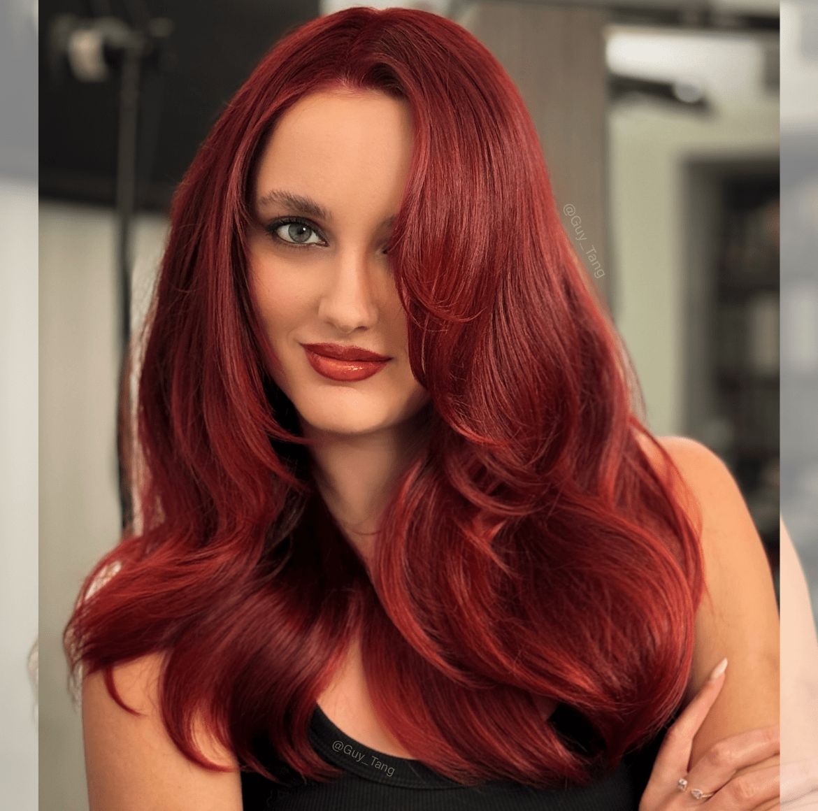 Woman with long, wavy fiery copper red hair and glossy finish, wearing a sleeveless top, posing in a salon with soft lighting.