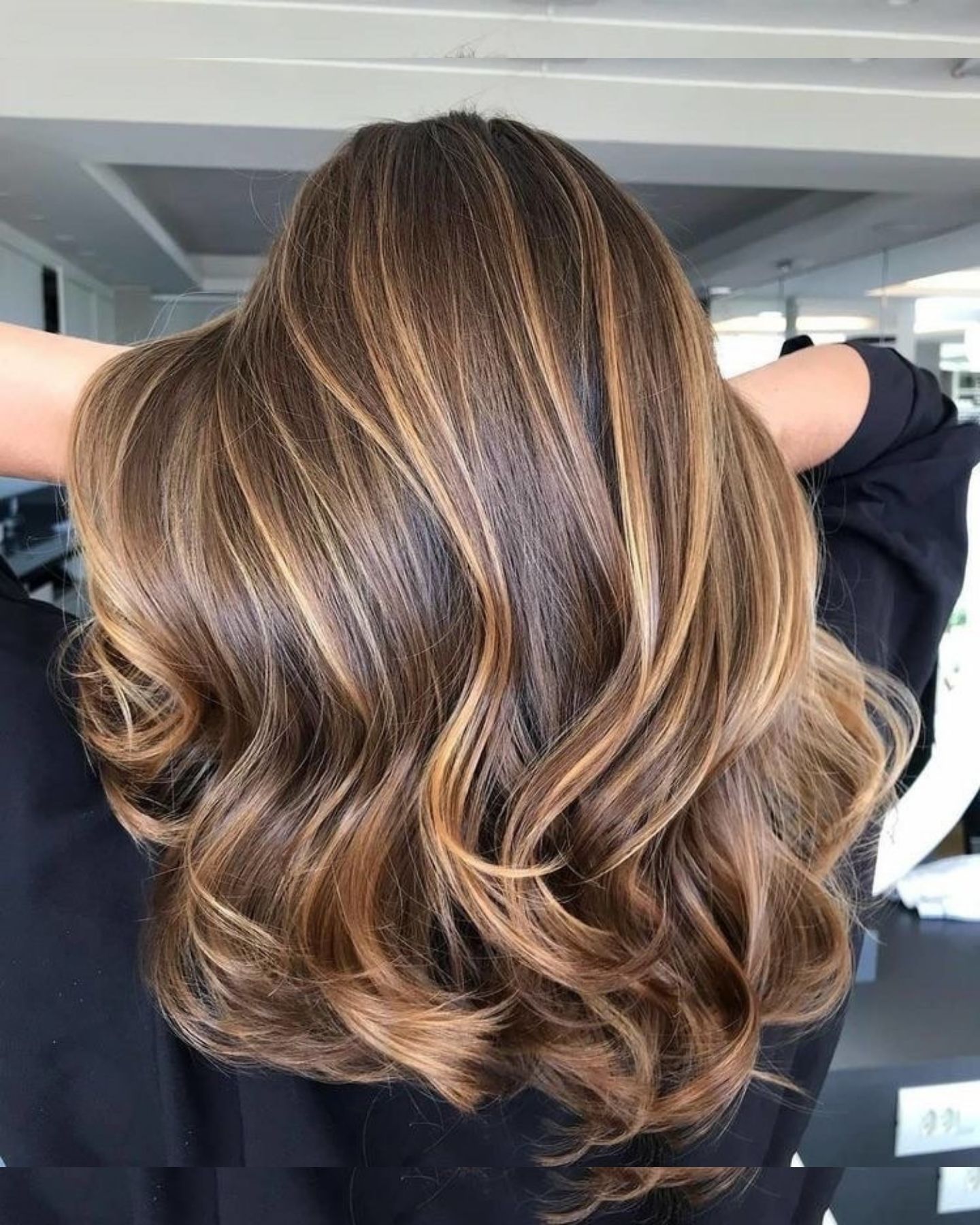 Rear view of a girl with medium-length layered hair featuring golden caramel highlights in a soft balayage blend, styled in voluminous waves with curled ends, wearing a black top in a modern indoor setting with clean lines and glass partitions.