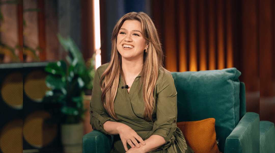 Kelly Clarkson with a round face shape sitting on a TV show sofa, smiling warmly with soft waves framing her full cheeks and soft jawline.