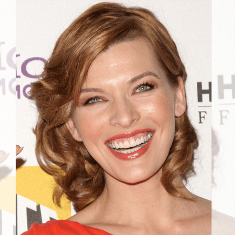 Milla Jovovich with a long face shape styled in a medium curly caramel updo, featuring soft waves and pinned-back layers for an elegant occasion-ready look.