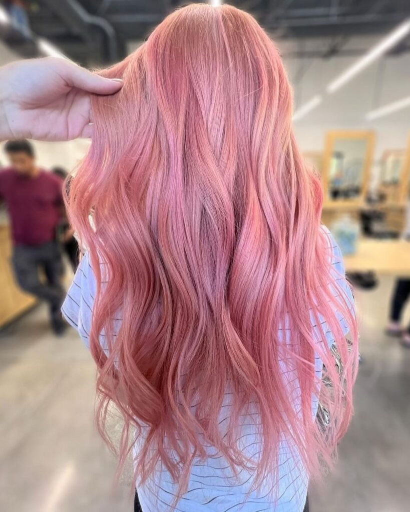 Back view of a girl with long, wavy pretty rose gold hair being adjusted by a hand, wearing a short-sleeved striped top in a modern salon setting with wooden accents, neutral flooring, and natural light filtering in.