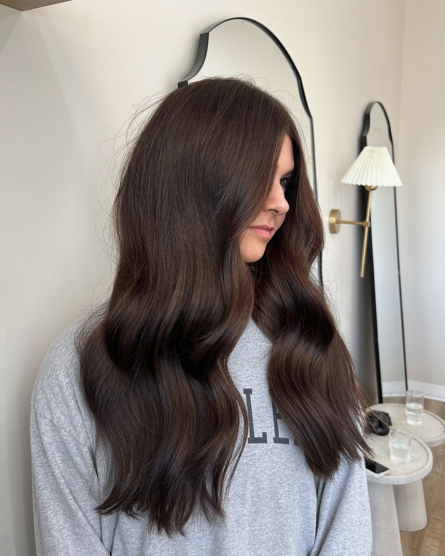 Girl with long, wavy rich chocolate brown hair styled in soft curls, wearing a grey sweatshirt with partial lettering, standing in a modern, light-filled room with minimalist furniture, including round side tables, a full-length arched mirror, and a pleated floor lamp.