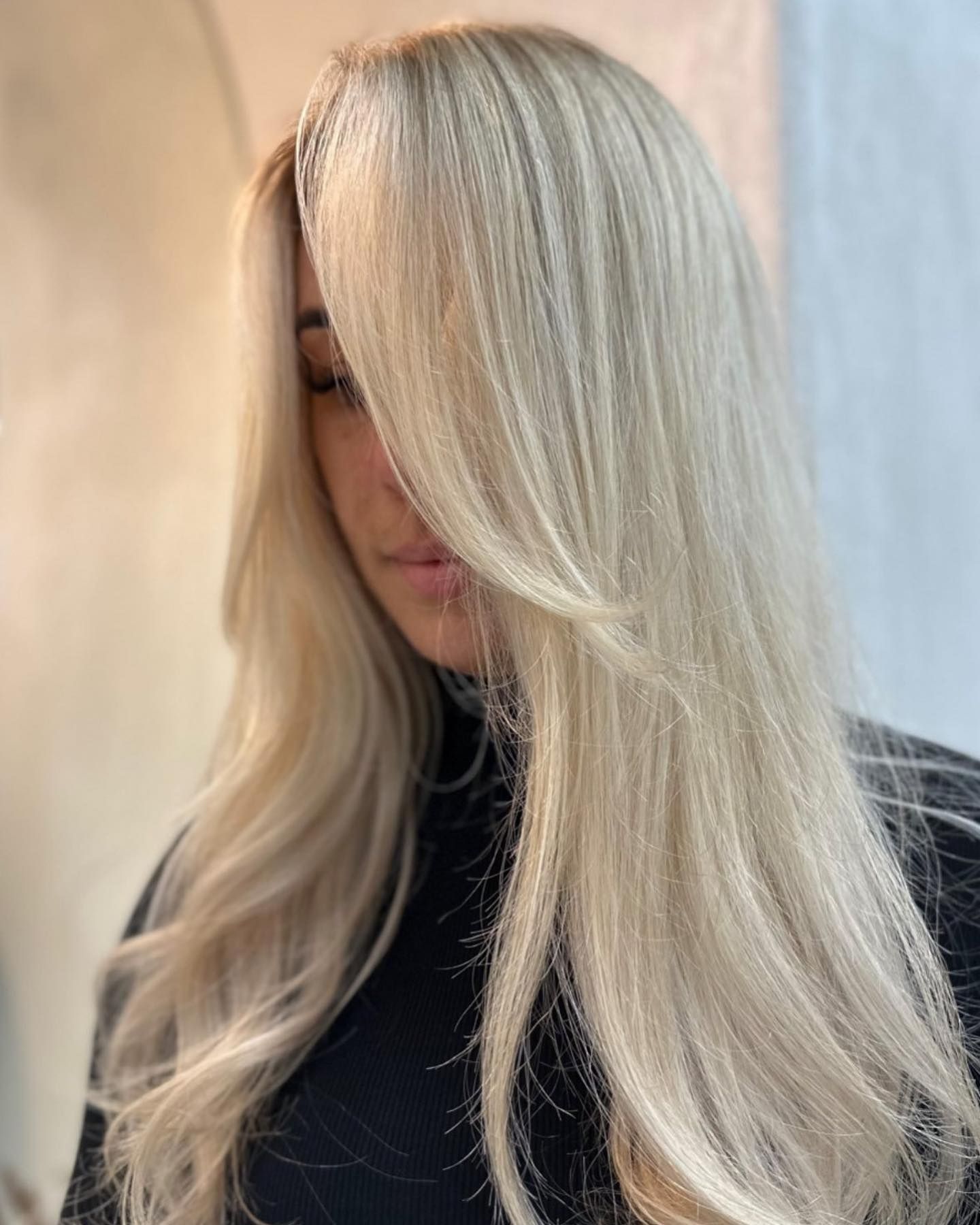 Girl with long, wavy sparkly champagne blonde hair cascading over her face and shoulders, wearing a fitted black round-neck knit top, standing in front of a softly lit neutral-toned background with a curtain or blinds filtering daylight.