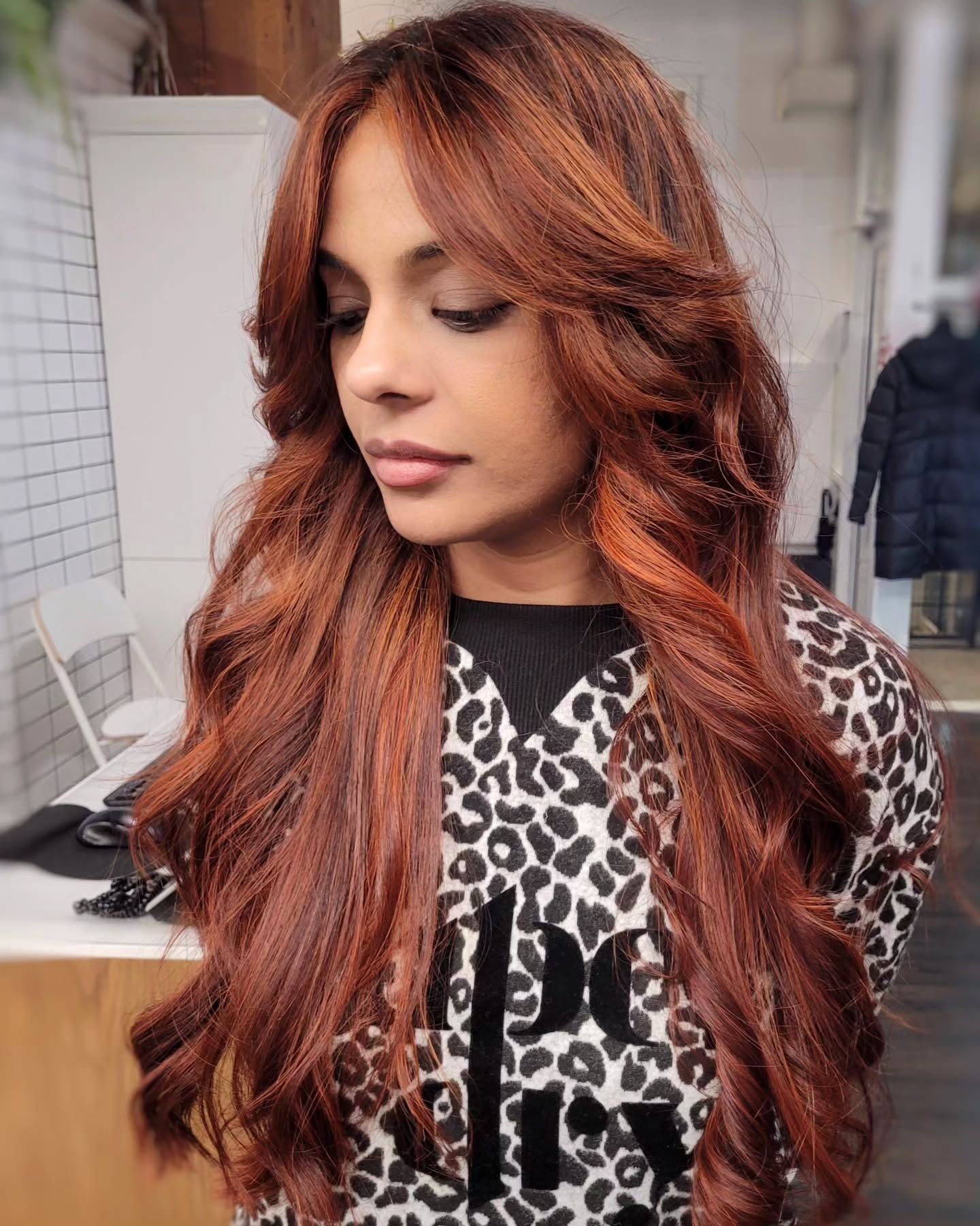 Girl with long, wavy spicy warm auburn hair styled in voluminous layers, wearing a white leopard print top, seen from the back while looking over her shoulder in a well-lit indoor setting with a blurred background showing a monitor and a coat rack.