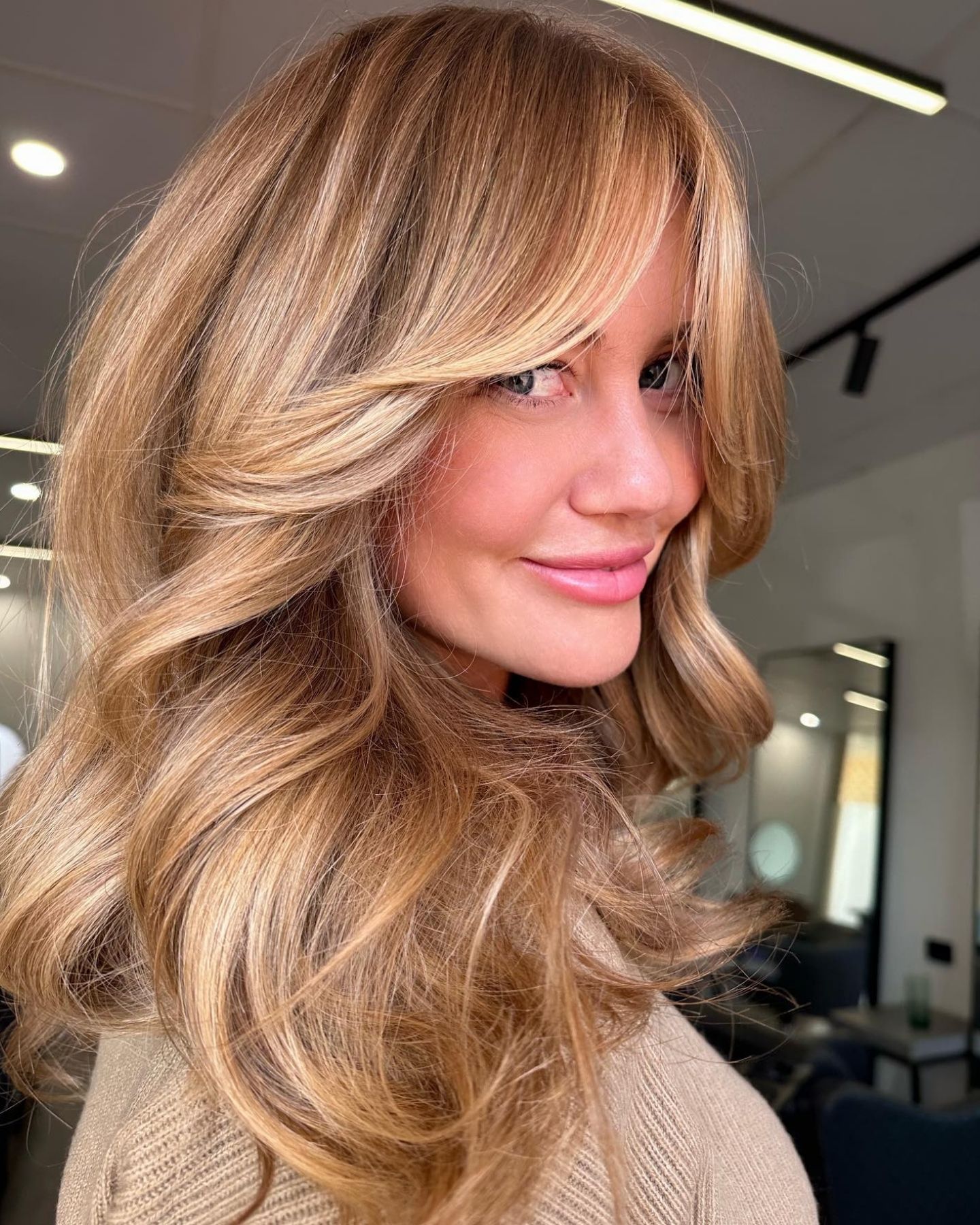 Girl with long, voluminous sunny honey balayage hair styled in soft waves, wearing a neutral knit sweater, in a modern indoor setting with minimalistic design elements and soft lighting reflecting off a large mirror.