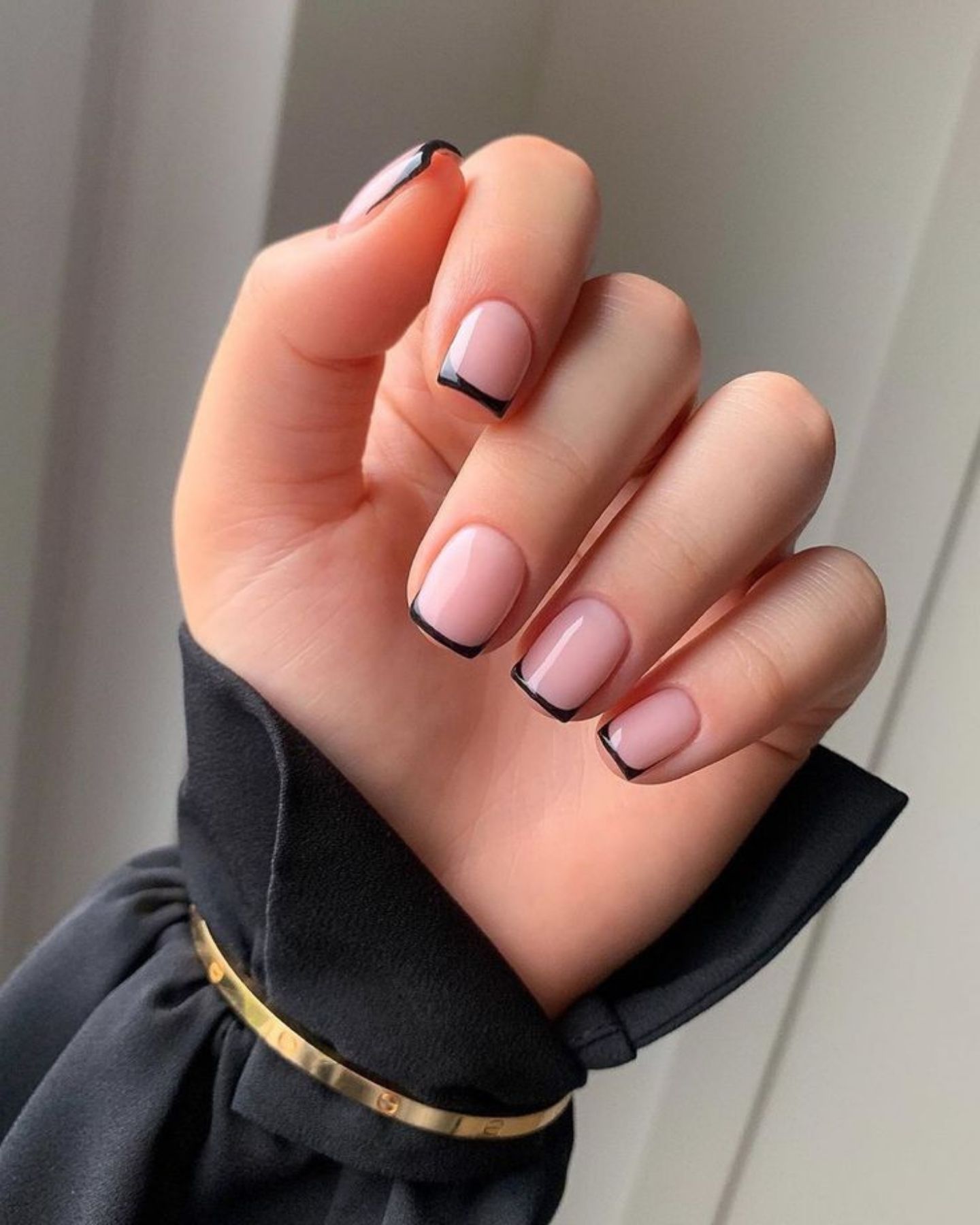 Short nails with black micro French tips and pale pink base, worn with a dark sleeve and subtle gold wrist accessory