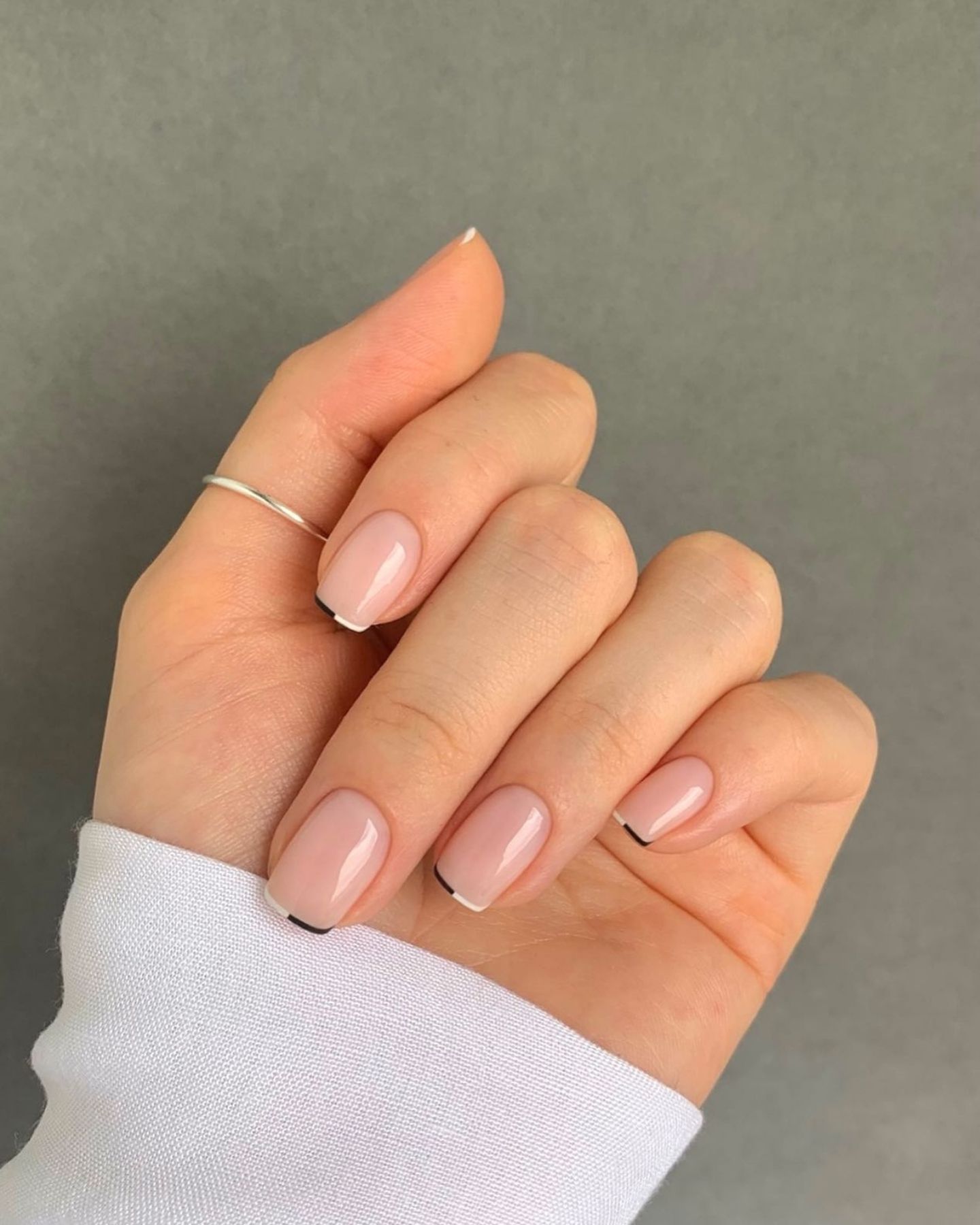 Black and white micro French nails on glossy nude base, with a silver ring on index finger and a white sleeve in a minimal backdrop.