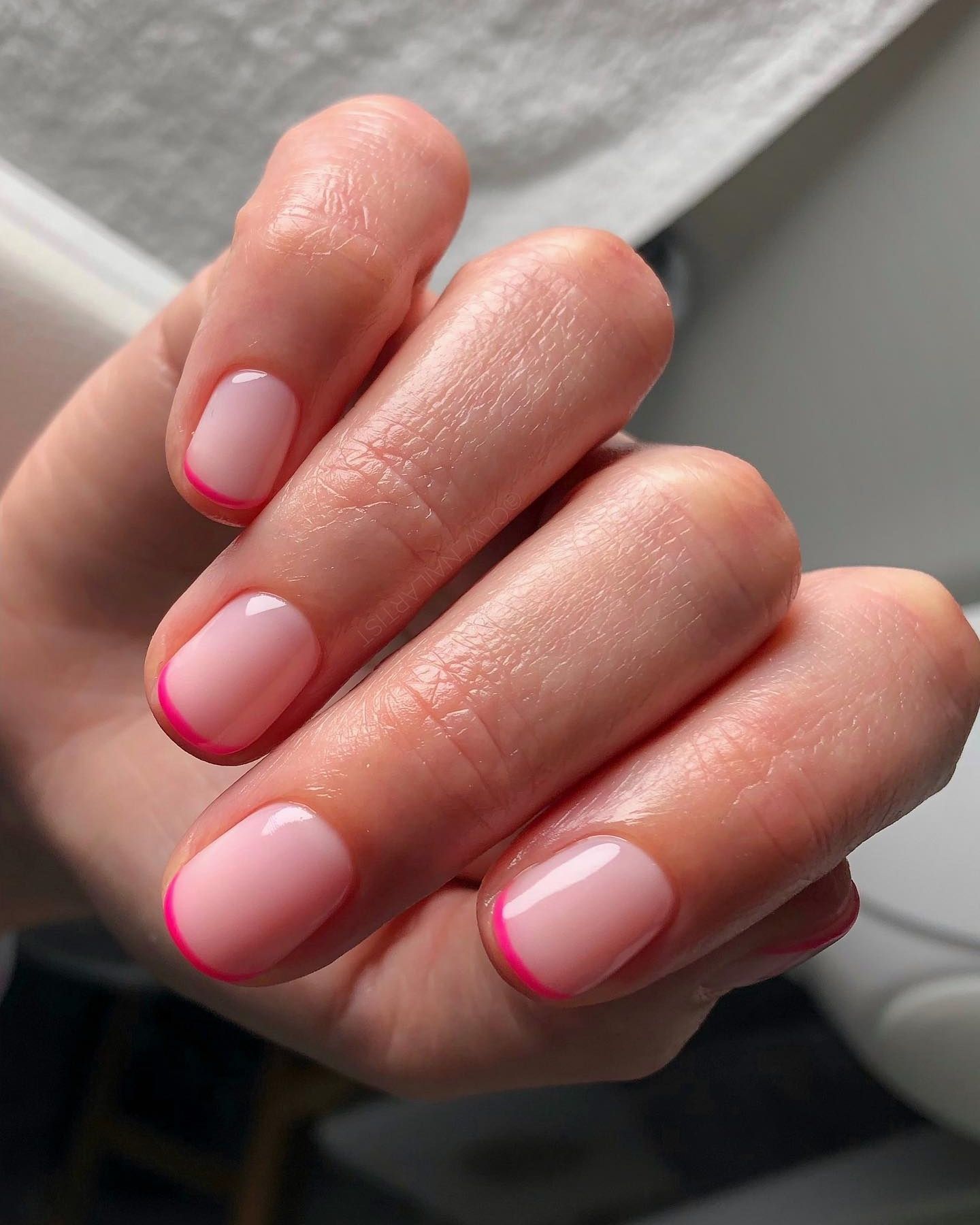 Bright pink micro French nails with a pale pink gradient base and accent nail, set against a neutral salon-like background.