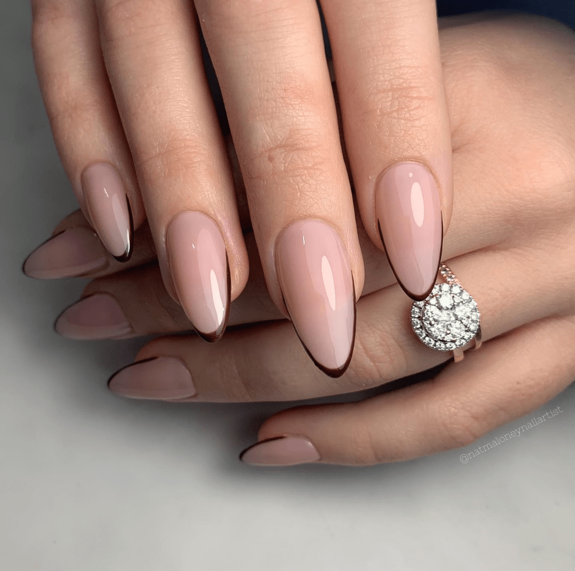 Brown micro French tips on almond-shaped nails with a light pink glossy base and gold diamond ring on the ring finger.
