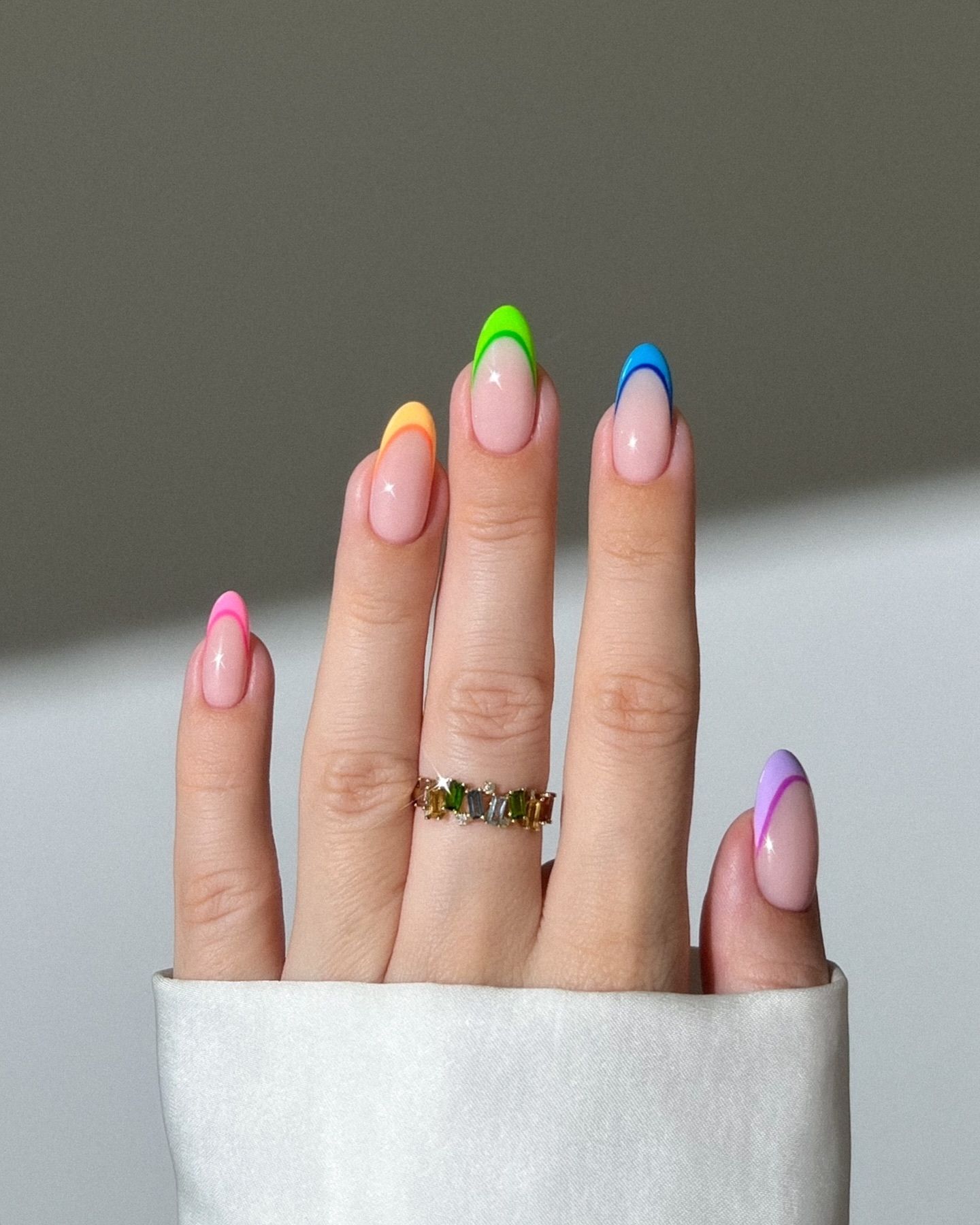 Almond-shaped nails with a colorful French tip gradient, paired with a gemstone ring, set against a grey background