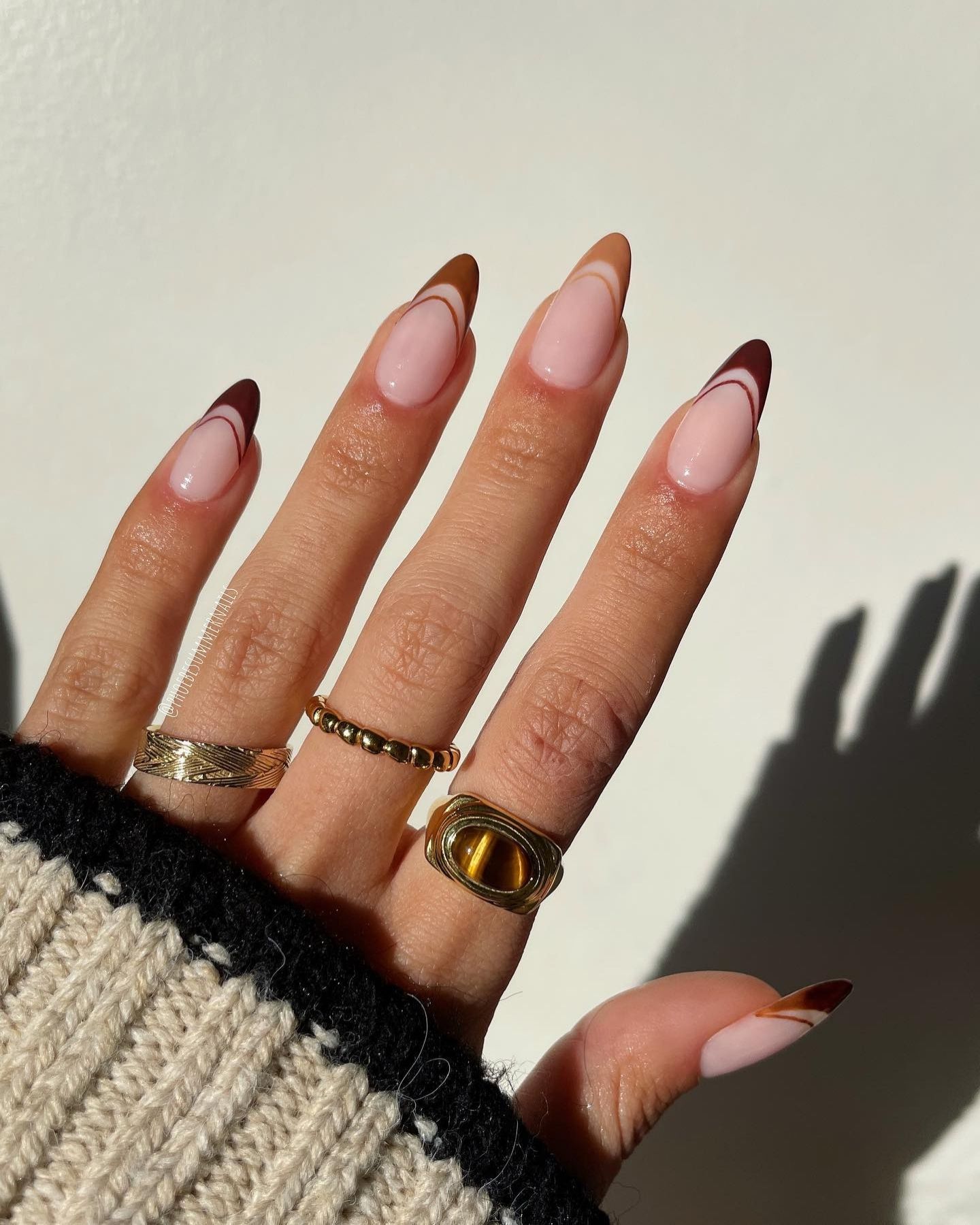 Stiletto-shaped nails with a Double French tip design, featuring a pastel pink base transitioning into a brownish hue at the tips. The nails are adorned with gold bands on the thumb, index, and middle fingers. The ring finger showcases a prominent gold ring with a tiger's eye stone set in a raised bezel. The hand is dressed in a knitted garment with black edges, and a soft shadow of the hand is cast onto the background.