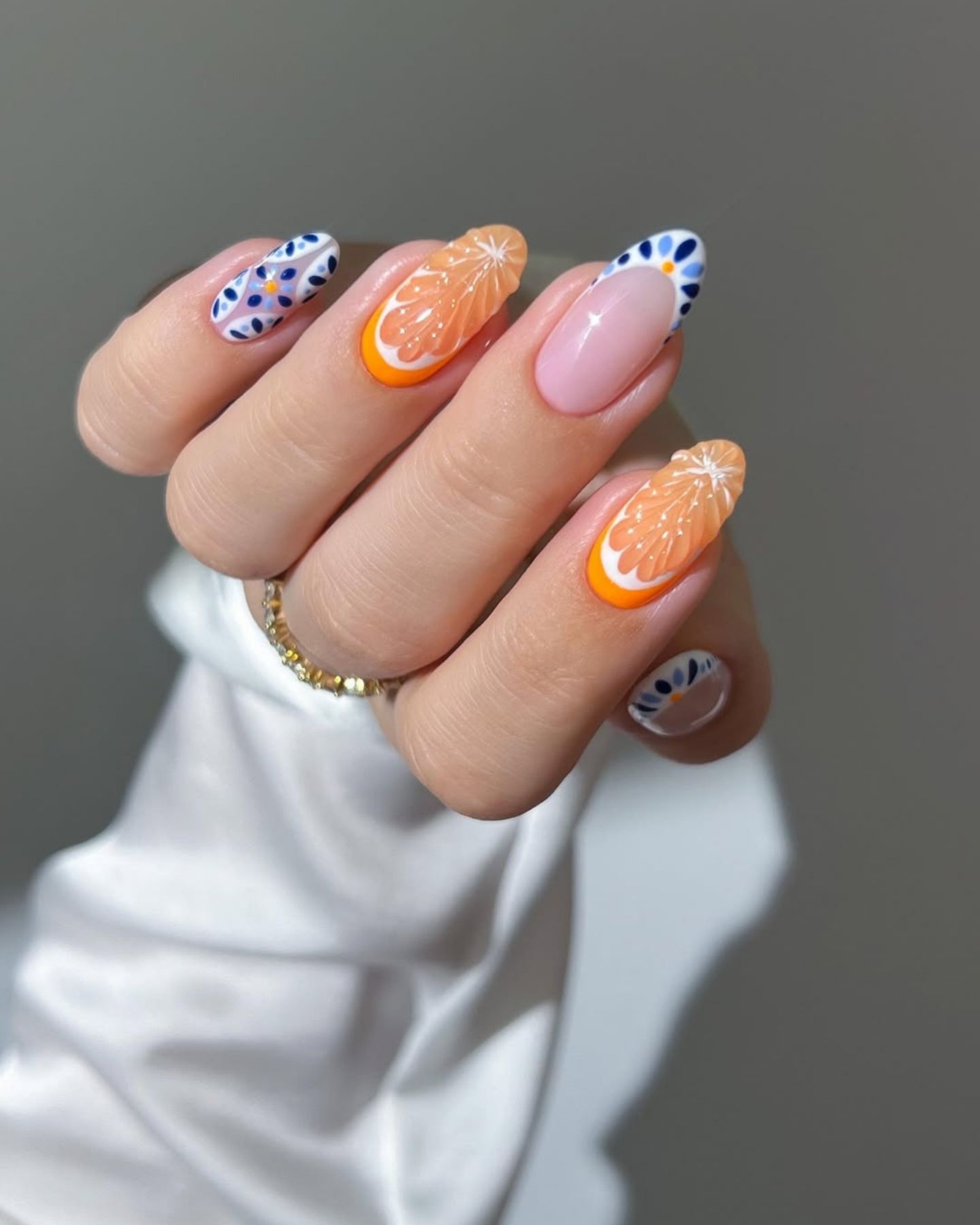 Hand with multicolored Euro summer nail art featuring pink ombré, citrus slice, floral orange, and geometric blue designs on a white satin background.