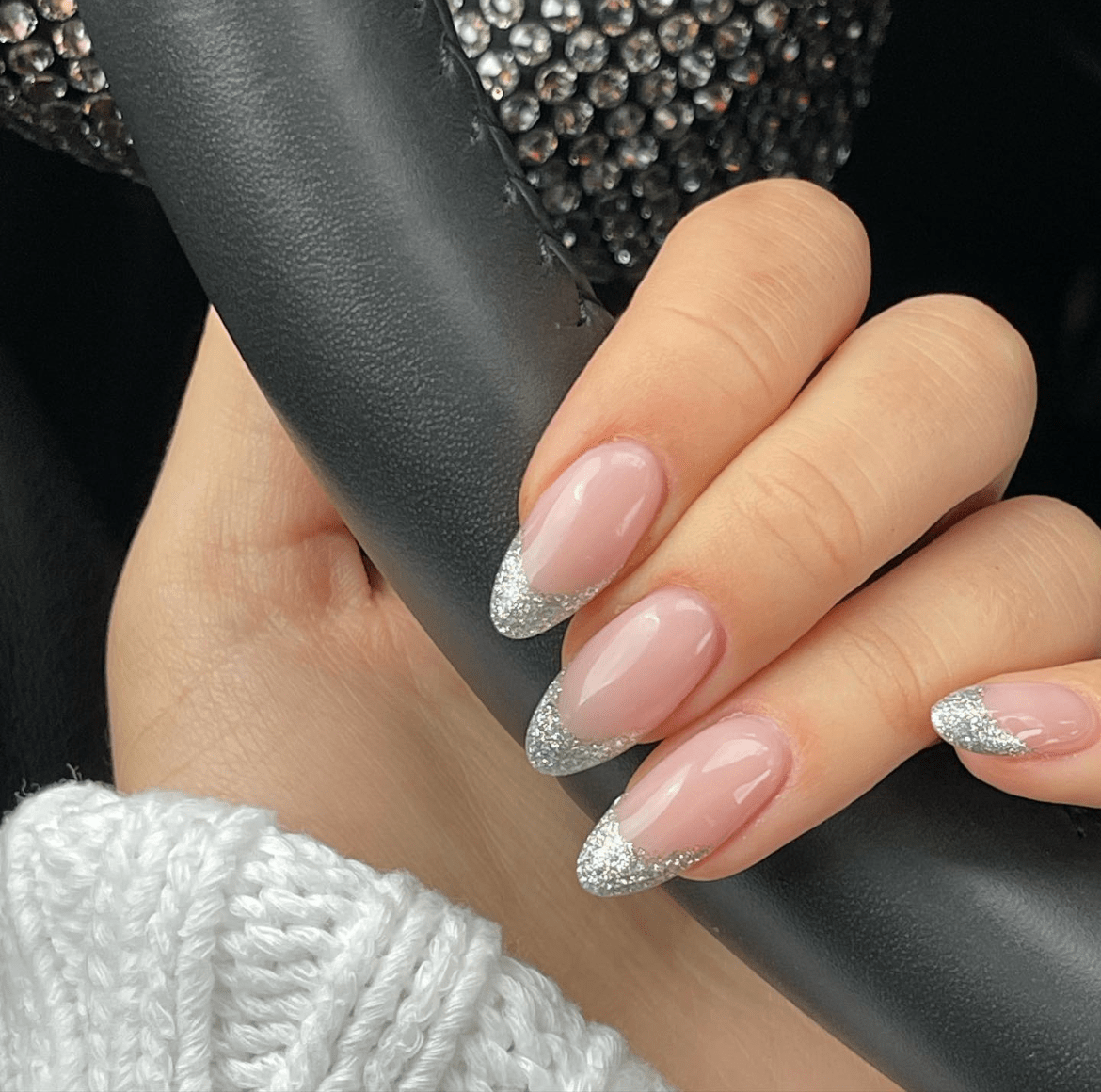 Glossy French tip nails with soft nude base and silver glitter tips, paired with knit sweater and black textured surface