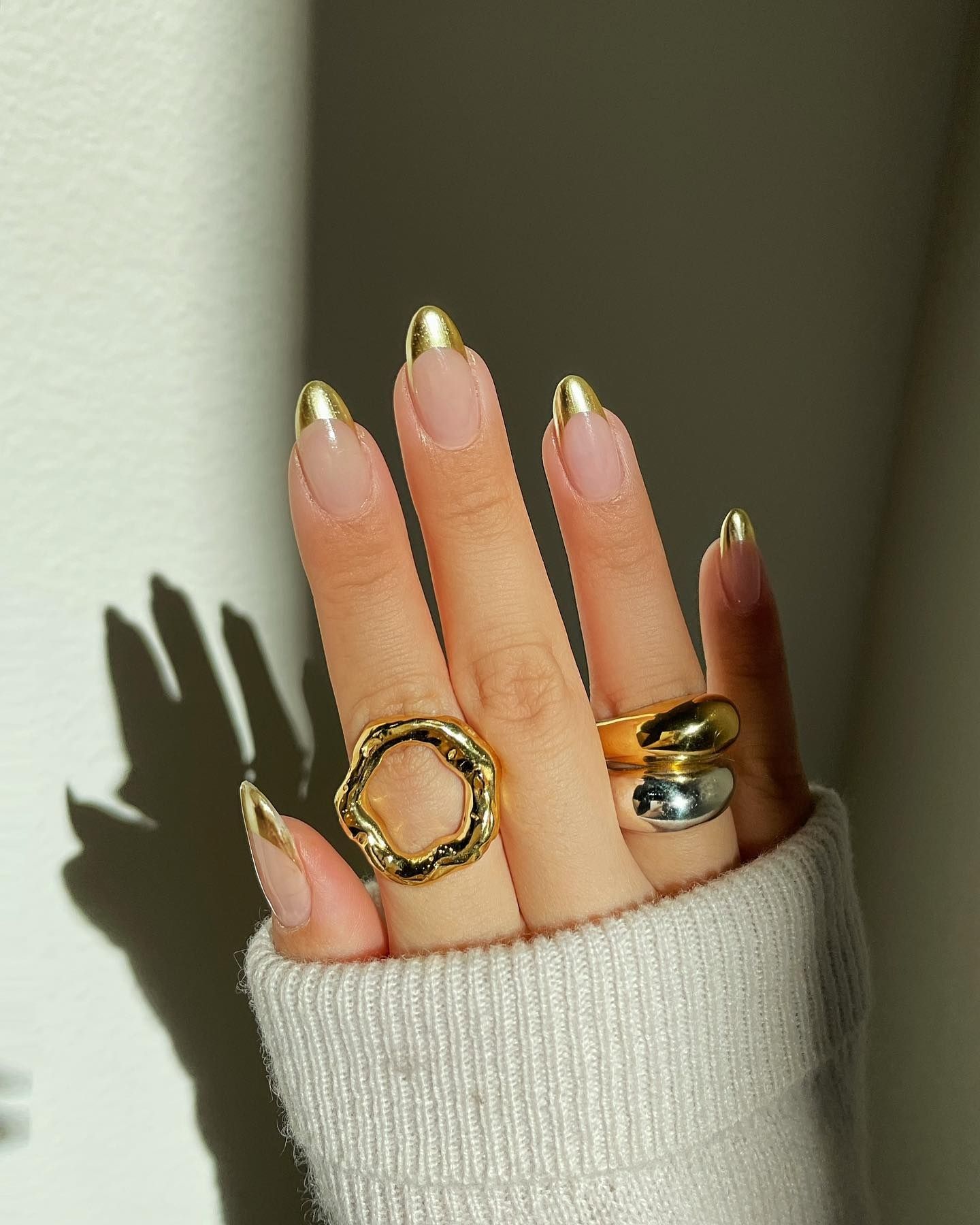 Gold French tip nails in stiletto shape with clear base, paired with gold and silver rings and ribbed knit sleeve