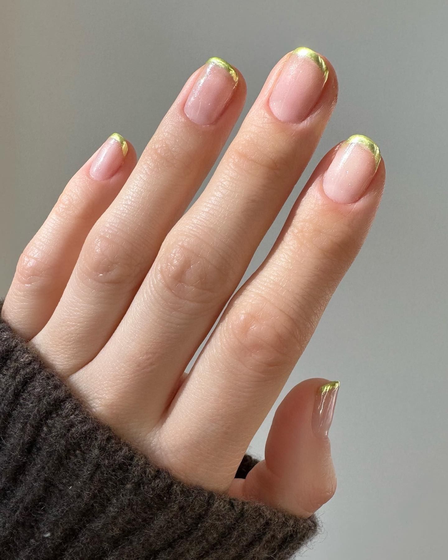 Gold micro French tips on pale pink nails, with fingers resting against ribbed charcoal knit fabric and a soft grey background.