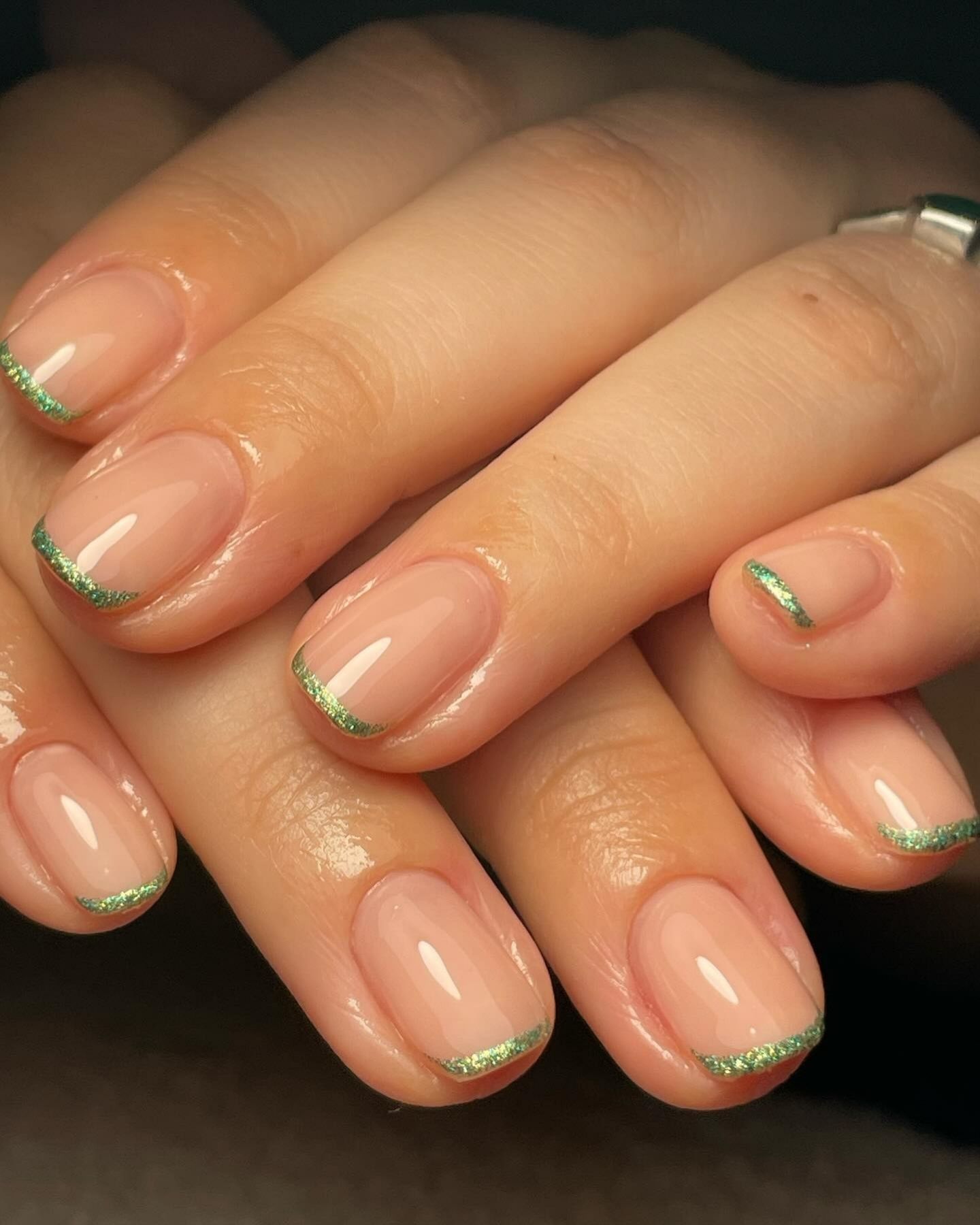 Close-up of manicured nails featuring a pale pink base with fine green glitter micro French tips, set against a dark background with soft indoor lighting.