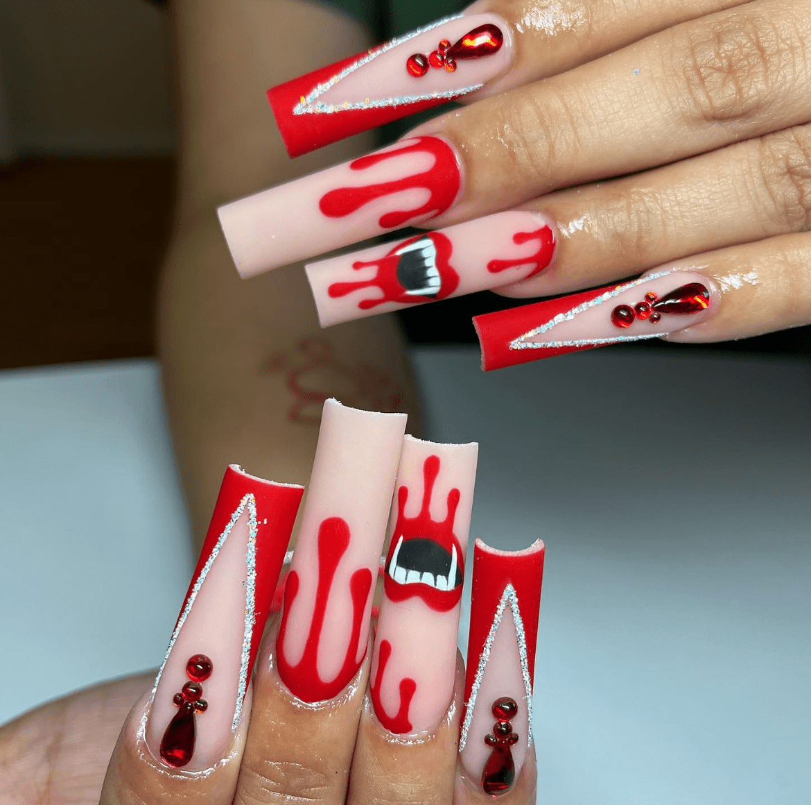 Halloween-inspired long acrylic nails with red glittery triangular tips, red gemstones, black triangle accents, and fang designs