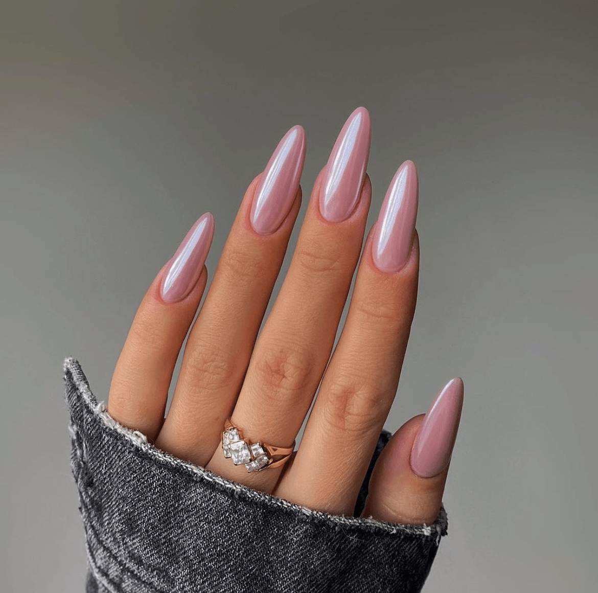 Almond-shaped nails painted in pearly light pink chrome, paired with a delicate gold ring with three tiny stones, resting on dark denim fabric.