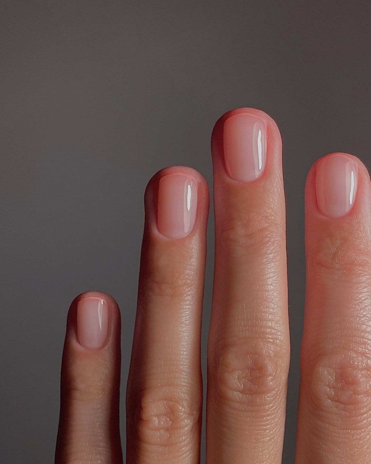 Close-up of four bent fingers with pale pink micro French tips on smooth skin, set against a solid grey background