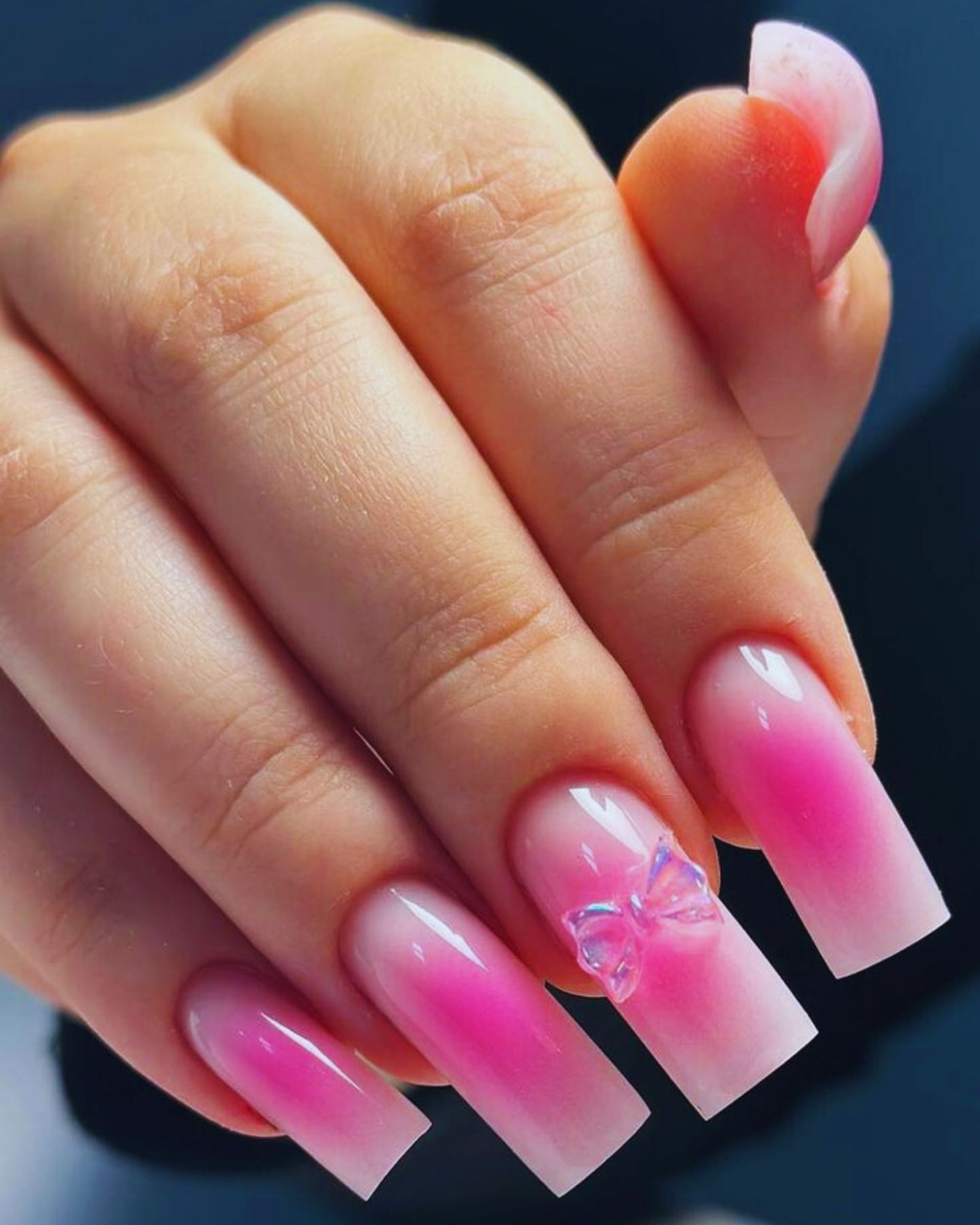 Close-up of long nails painted in a glossy gradient from dark pink tips to lighter pink bases, with a curved, stiletto-style index finger adorned with a clear bow design. The nails shine with a smooth, polished finish, showcasing a blend of soft elegance and bold creativity.