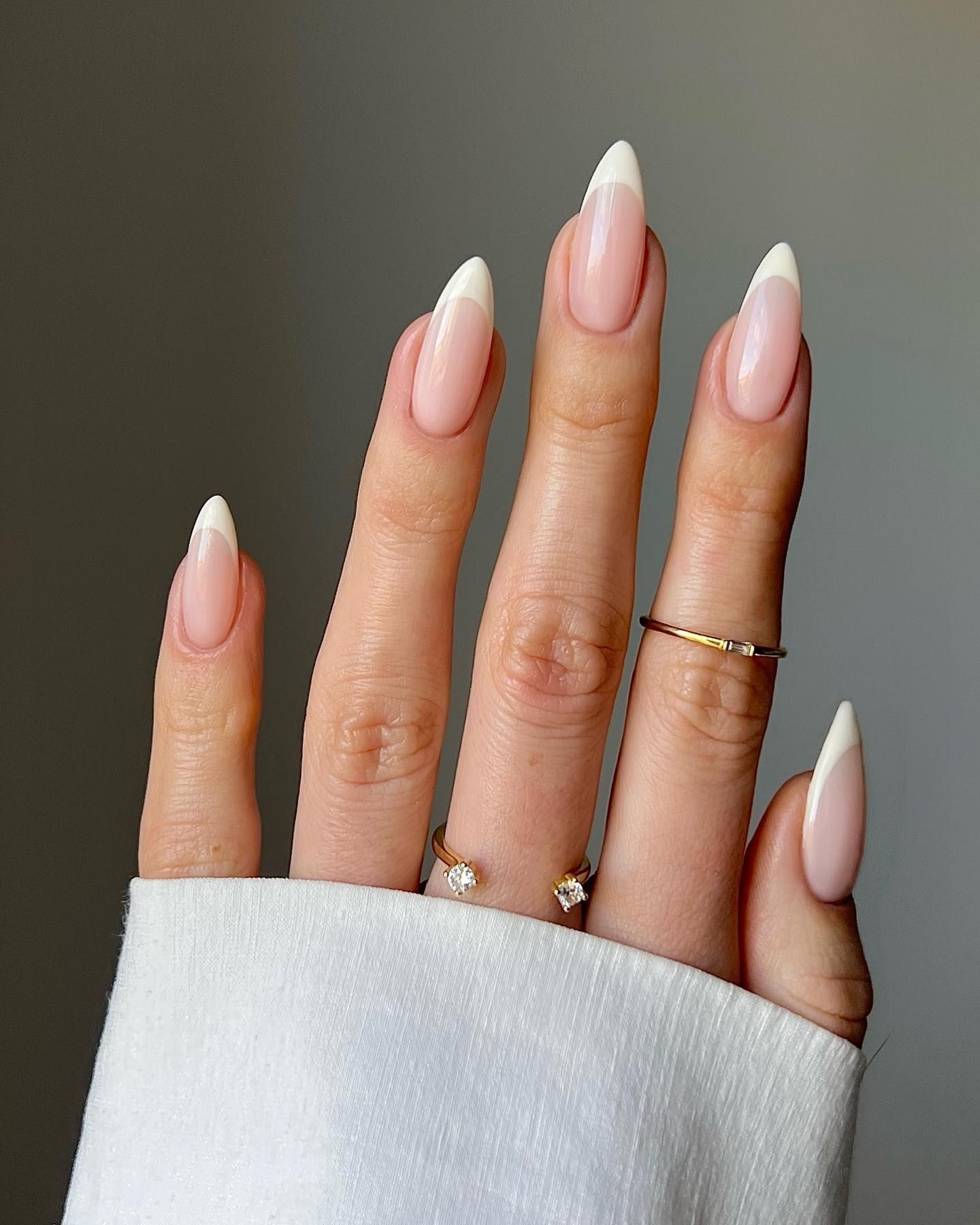 Close-up of glossy almond-shaped nails with pale pink polish, accented by three elegant gold rings with diamonds on a soft grey background