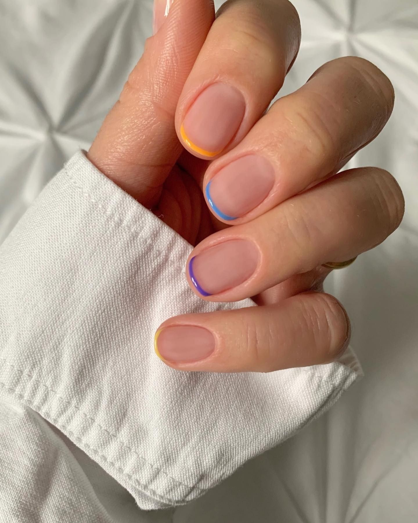 Hand with multicolored micro French nails featuring pastel bases and yellow tips, resting on a white textured bedsheet.
