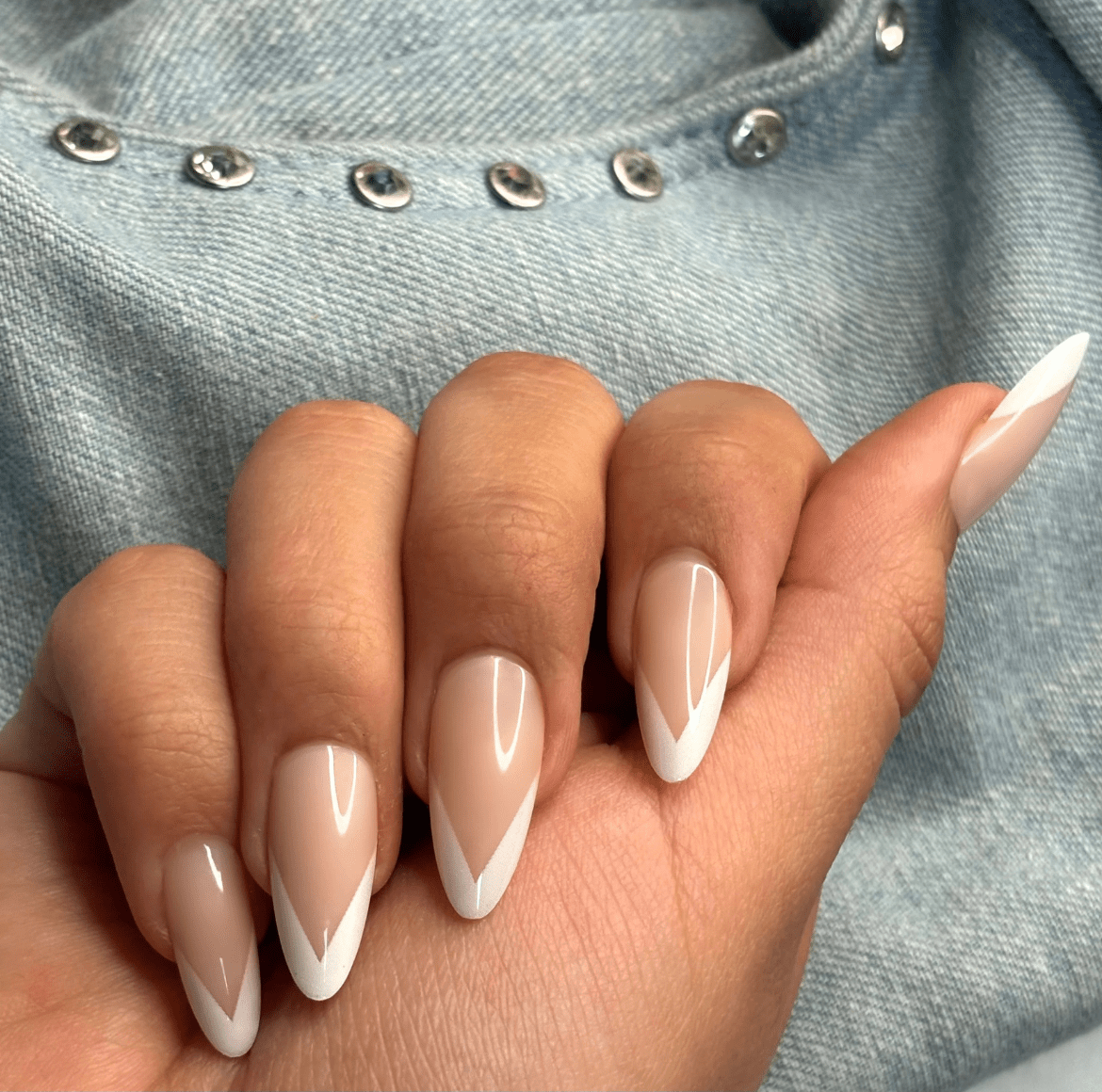 Natural V-shaped nails with glossy nude base and double white V tips against denim fabric with metallic buttons
