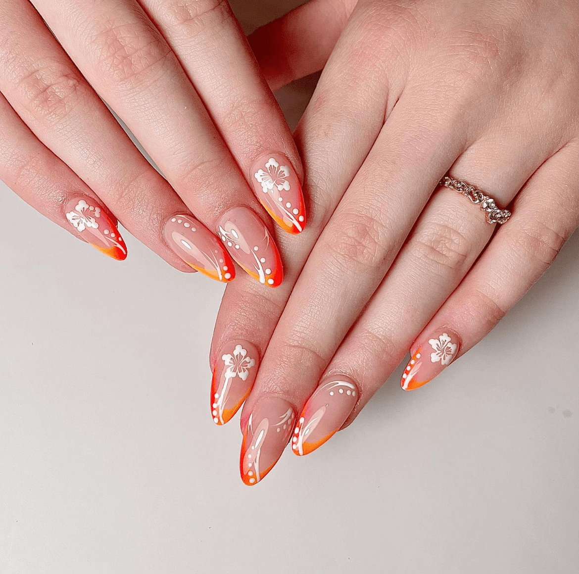 Two hands with stiletto-shaped nails featuring vibrant neon orange French tips that fade into a clear base. The nails are adorned with hand-painted white flowers, green leaves, and delicate swirling accents. A silver ring with gemstones sits on one finger, and the background is a soft, neutral tone that highlights the intricate nail design.