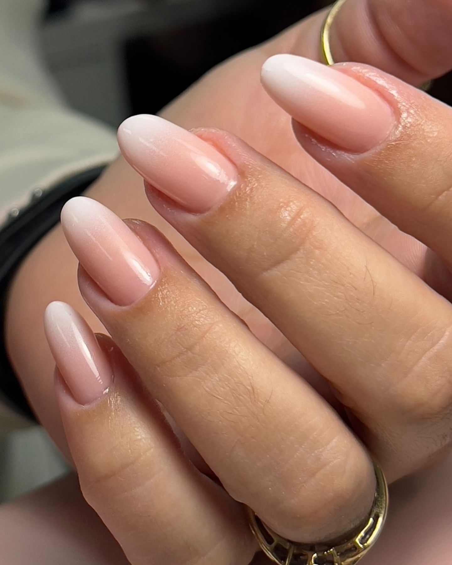 Almond-shaped Baby Boomer nails with a pink-to-white ombré gradient, worn on a well-groomed hand with a gold ring on the ring finger against a neutral blurred background.