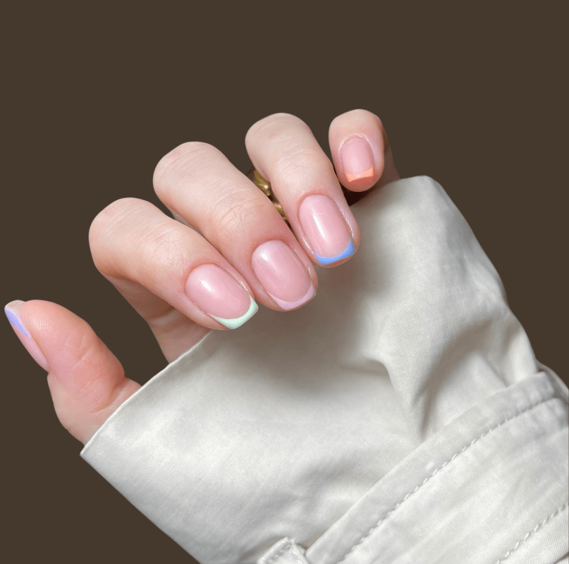 Hand with pastel micro French nails featuring alternating pink, blue, and orange tips over a clear light pink base, partially wrapped in a white cuff against a neutral brown background.