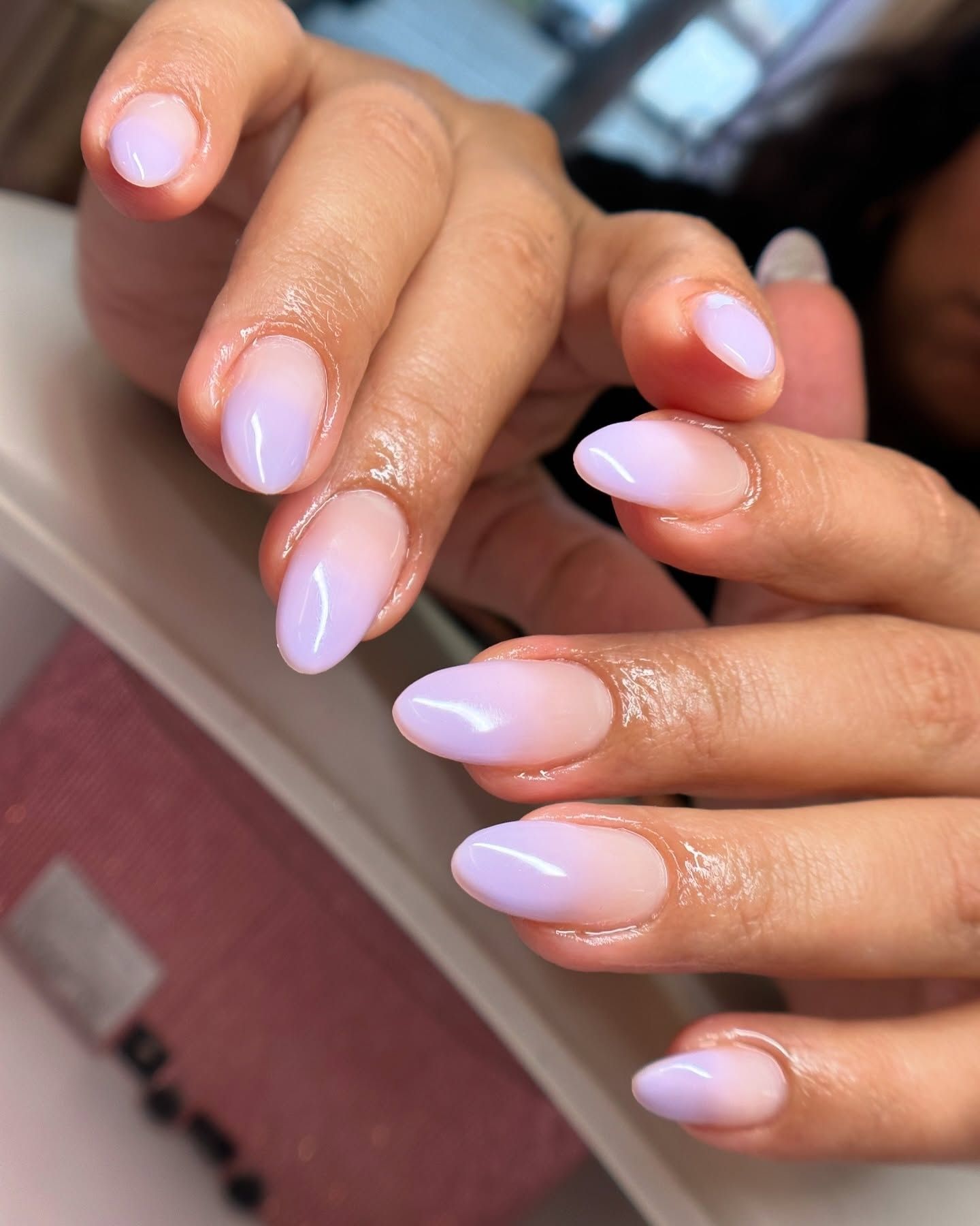 Almond-shaped gel nails with a glossy pastel ombré design blending soft pink and lavender, displayed on hands with a tan skin tone.