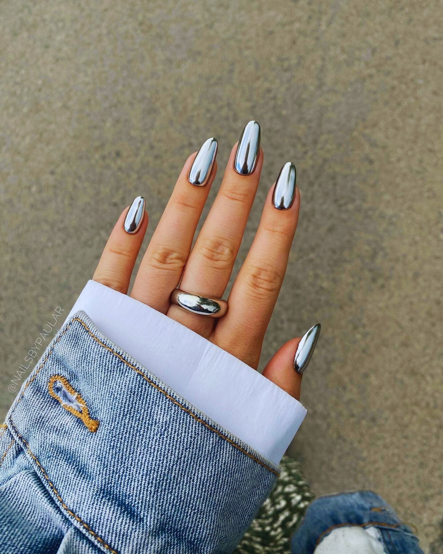 Close-up of a hand with sleek silver chrome almond-shaped nails, a silver band ring on the ring finger, and a portion of a light-wash denim jacket sleeve. The reflective nails contrast elegantly with the casual, modern aesthetic.
