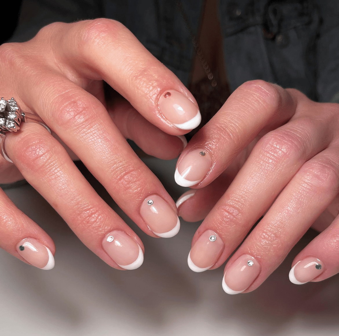Short French nails with a clear base and delicate gemstone accents, featuring varied nail lengths and a subtle white tip on the pinky.