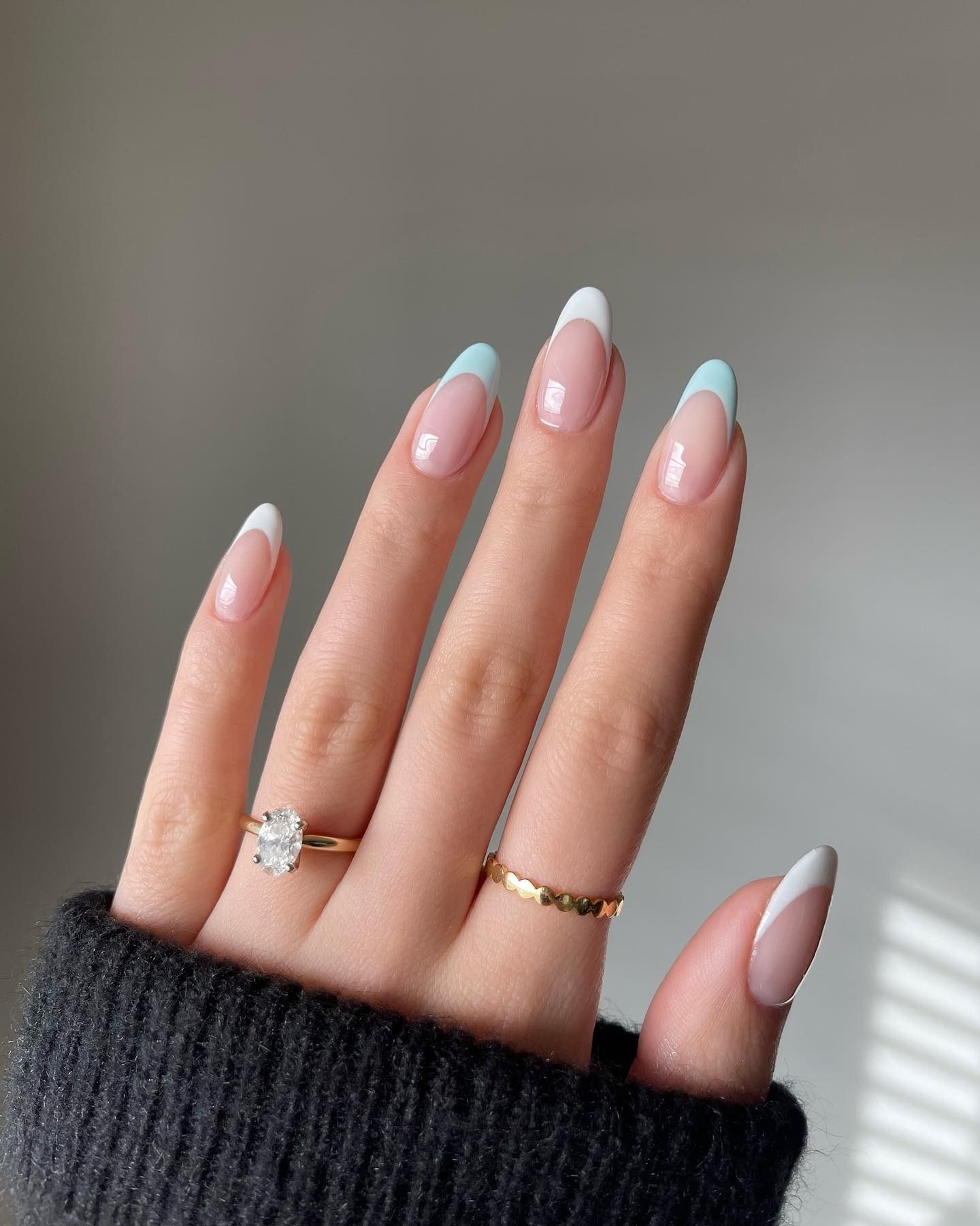 Close-up of almond-shaped nails with pastel blue French tips and a nude base, complemented by two rings on the ring and middle fingers — a solitaire diamond engagement ring and a whimsical gold heart-link ring. The soft, spring-inspired nail design highlights the elegant curves of the nails.
