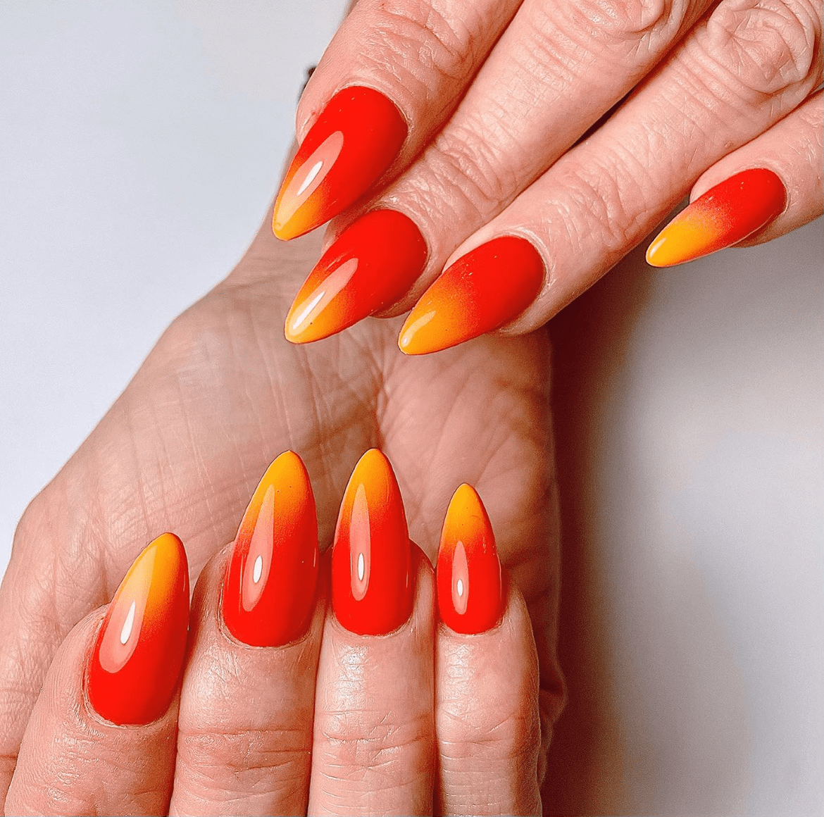 Hands with long almond-shaped nails featuring a vibrant sunset ombré design blending bright yellow tips into bold red bases, set against a plain white background