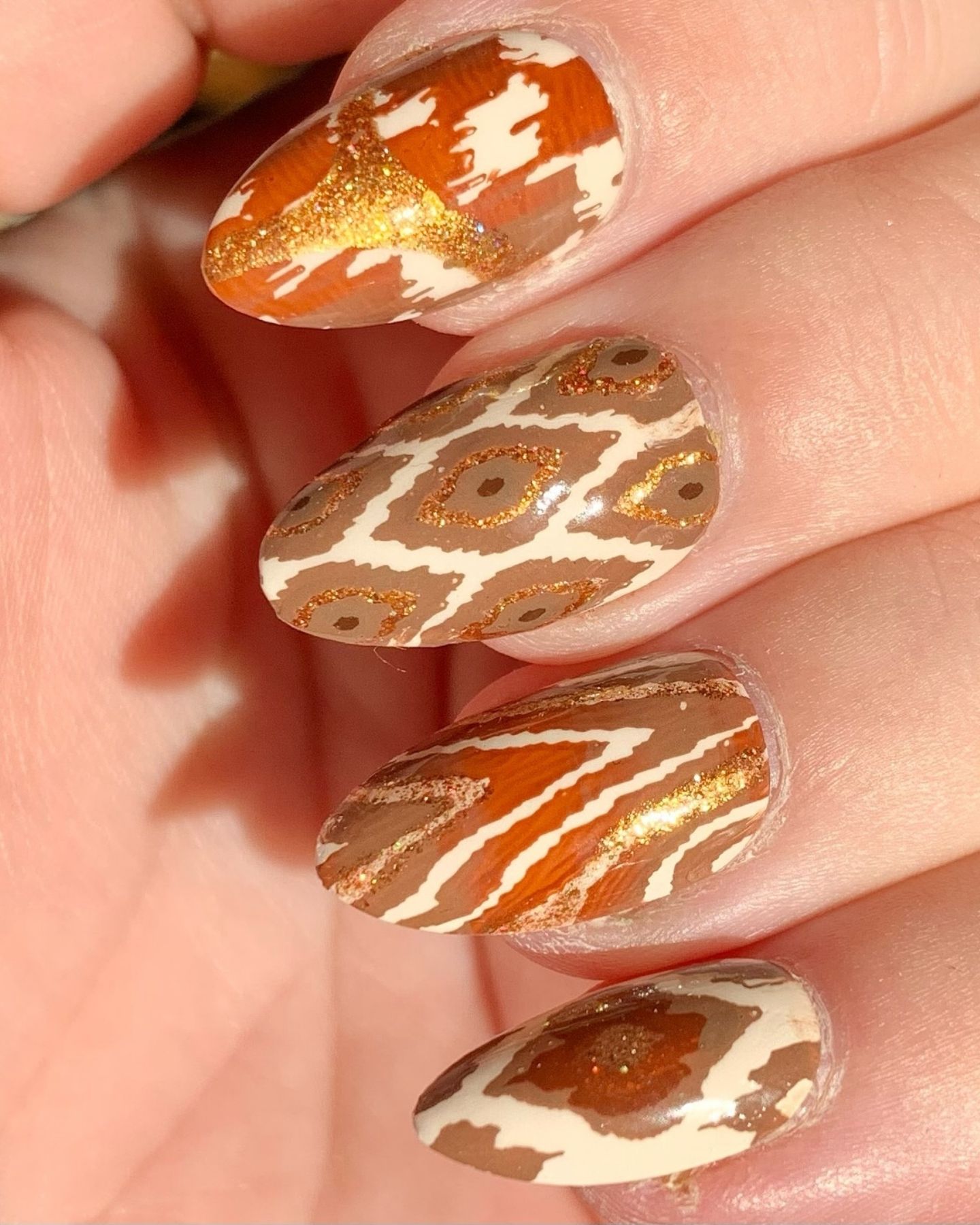 Close-up of Textile Pattern Nail Art on almond-shaped nails, featuring a mix of earthy tones with giraffe-inspired prints, abstract brown splashes, orange strokes, and gold glitter details for a bold, nature-themed look.