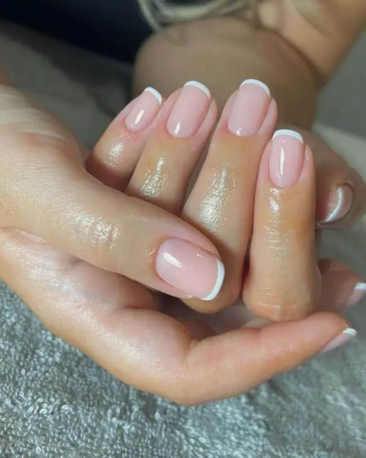 Close-up of hands with thin white French tips on glossy pink nails, gently clasped over a soft grey textured surface