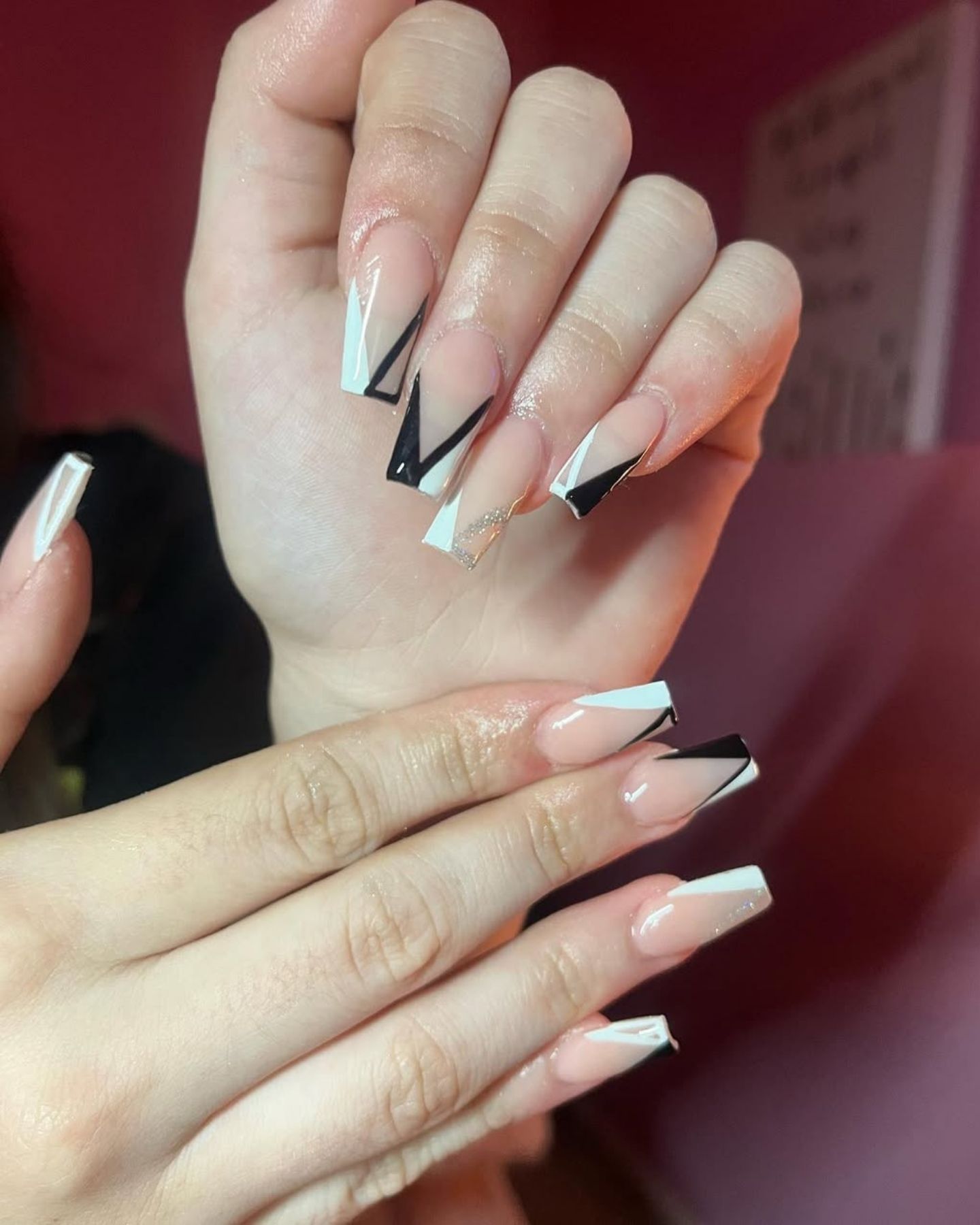 Long clear stiletto nails with glossy V-French tips, featuring sharp black and white geometric designs accented with fine glitter; a second hand with shorter nails and minimalist white lines complements the look.