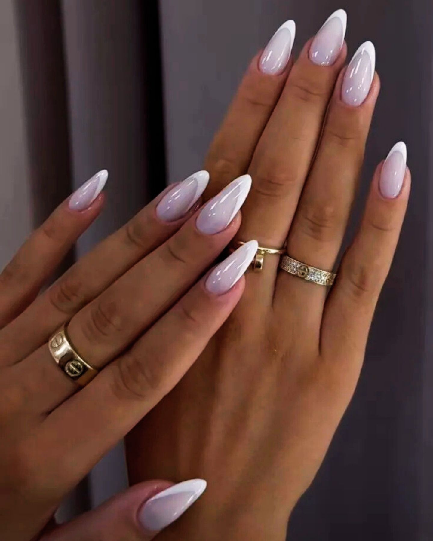 White French tip nails in almond shape with pastel pink gradient and glossy finish, accented by gold diamond rings