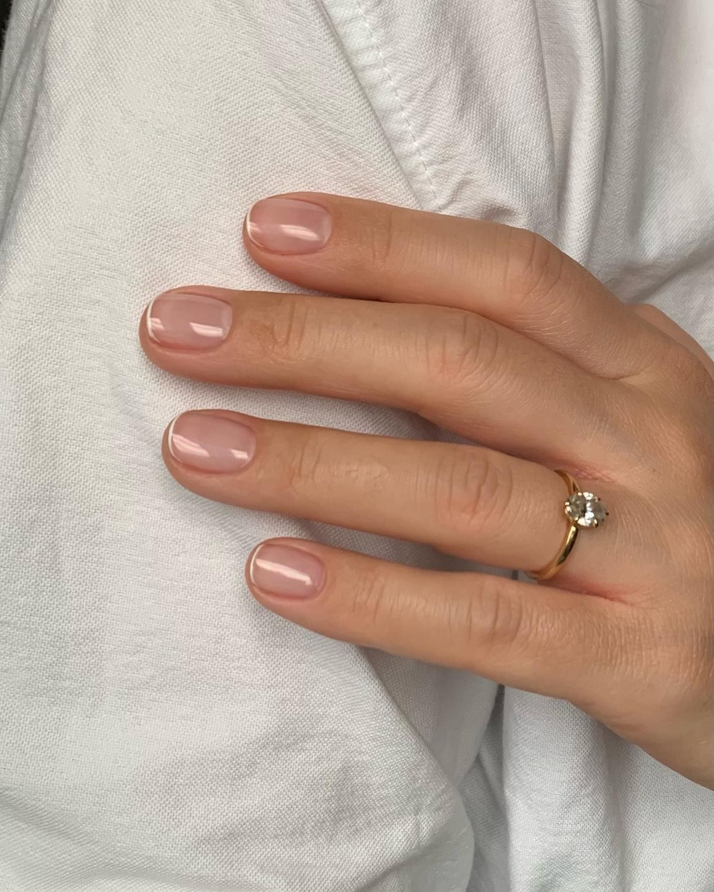 Hand with natural pink-to-peach gradient nails featuring white micro French tips, wearing a gold engagement ring with a round-cut diamond, resting on soft white fabric.