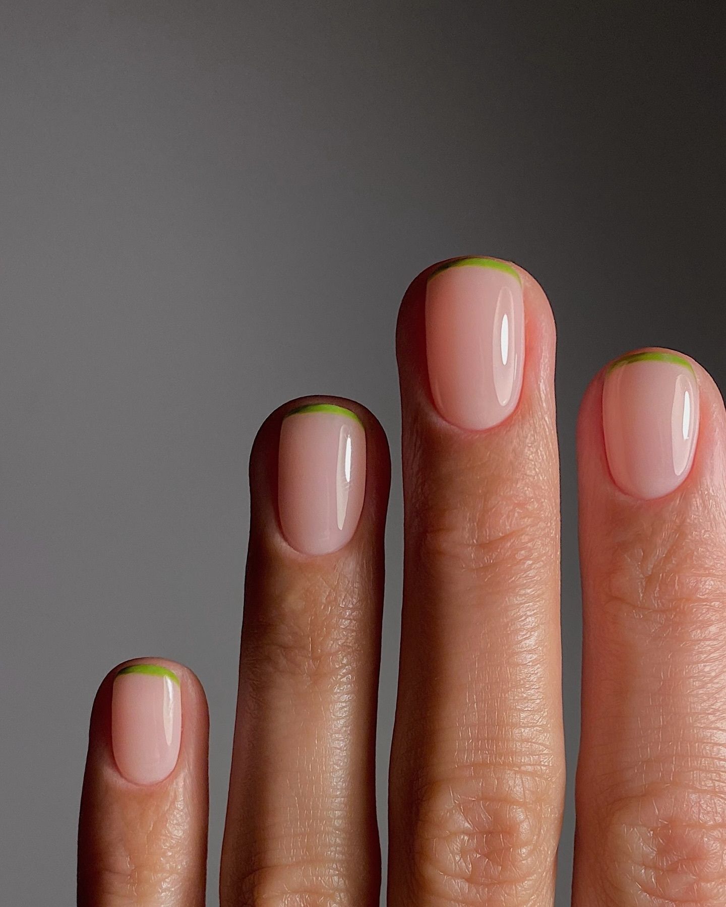 Pale pink nails with minimalist green tips and abstract green stripes, shown on a close-up hand against a neutral gray background