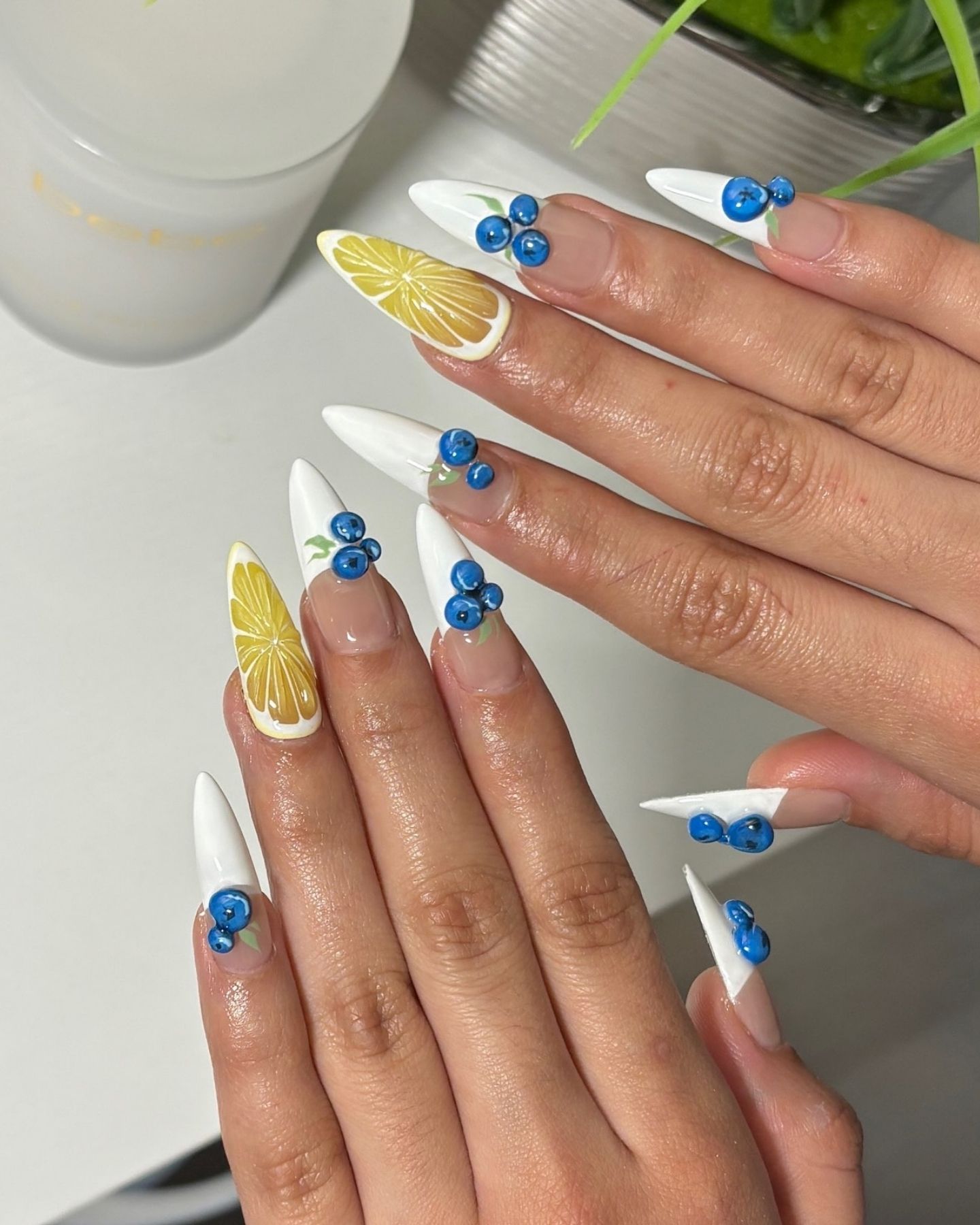 Pointed white nails with hand-painted blueberry and lemon slice designs, accented with green leaves and styled in a refreshing summer theme