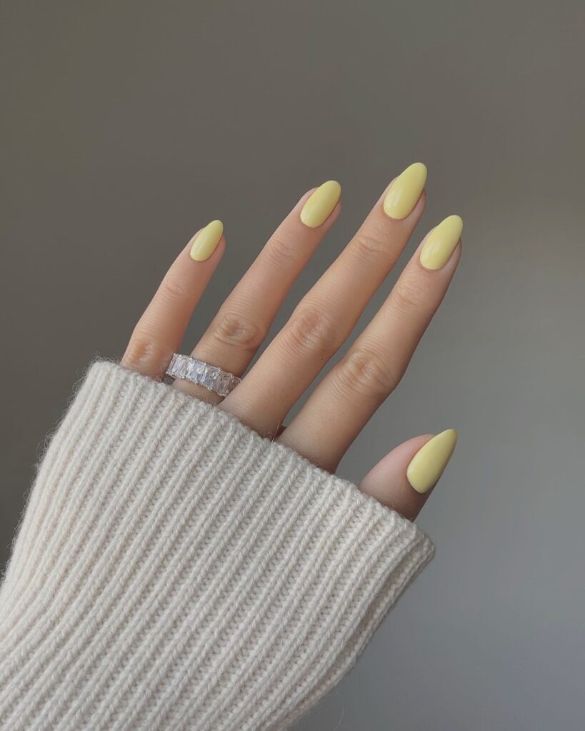 Almond-shaped pastel yellow nails with a glossy finish, styled with a silver ring and cozy off-white knit sleeve.