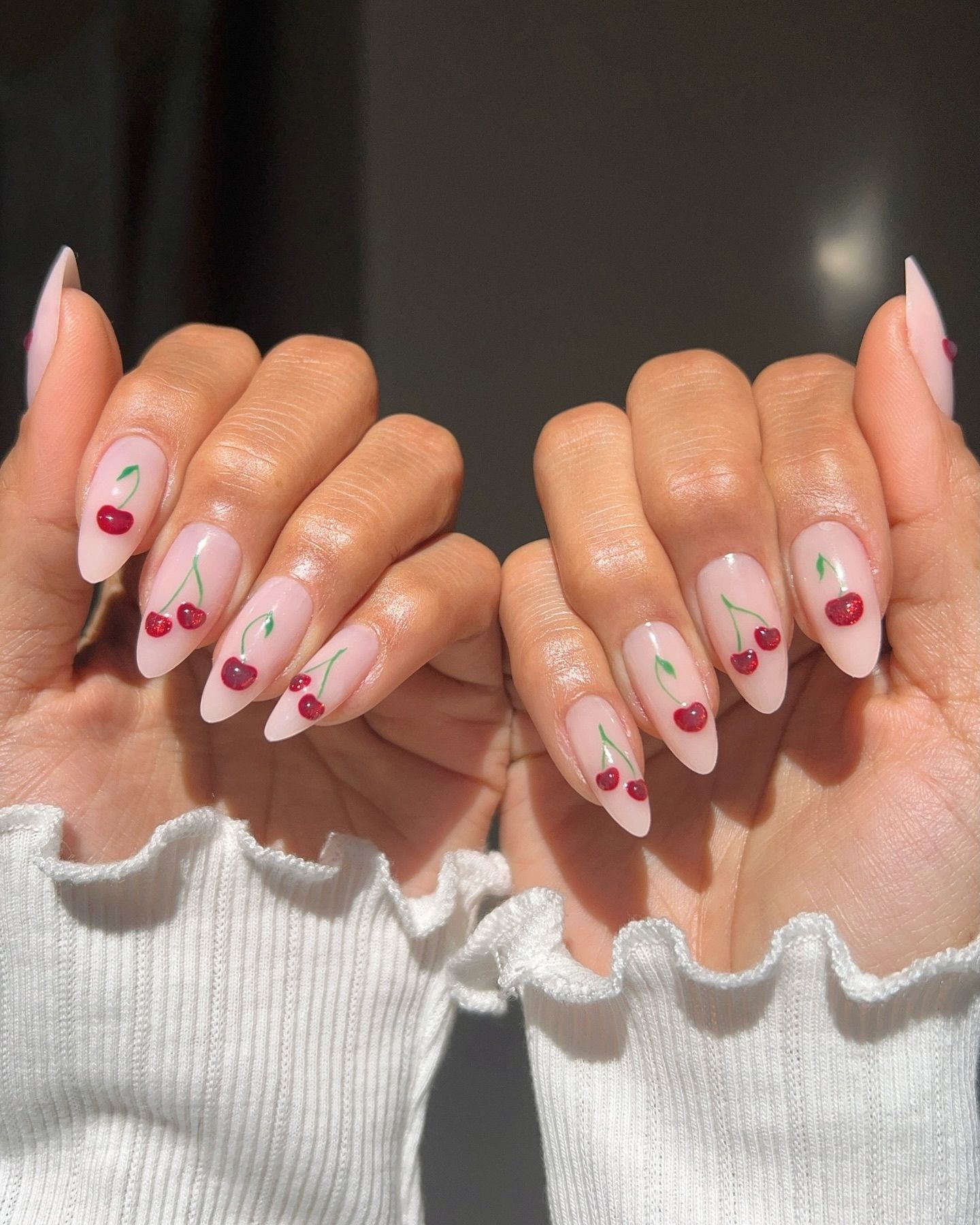 Almond-shaped nails with translucent pink polish and detailed red cherry designs, accented by green stems and leaves, styled with soft white cuffs