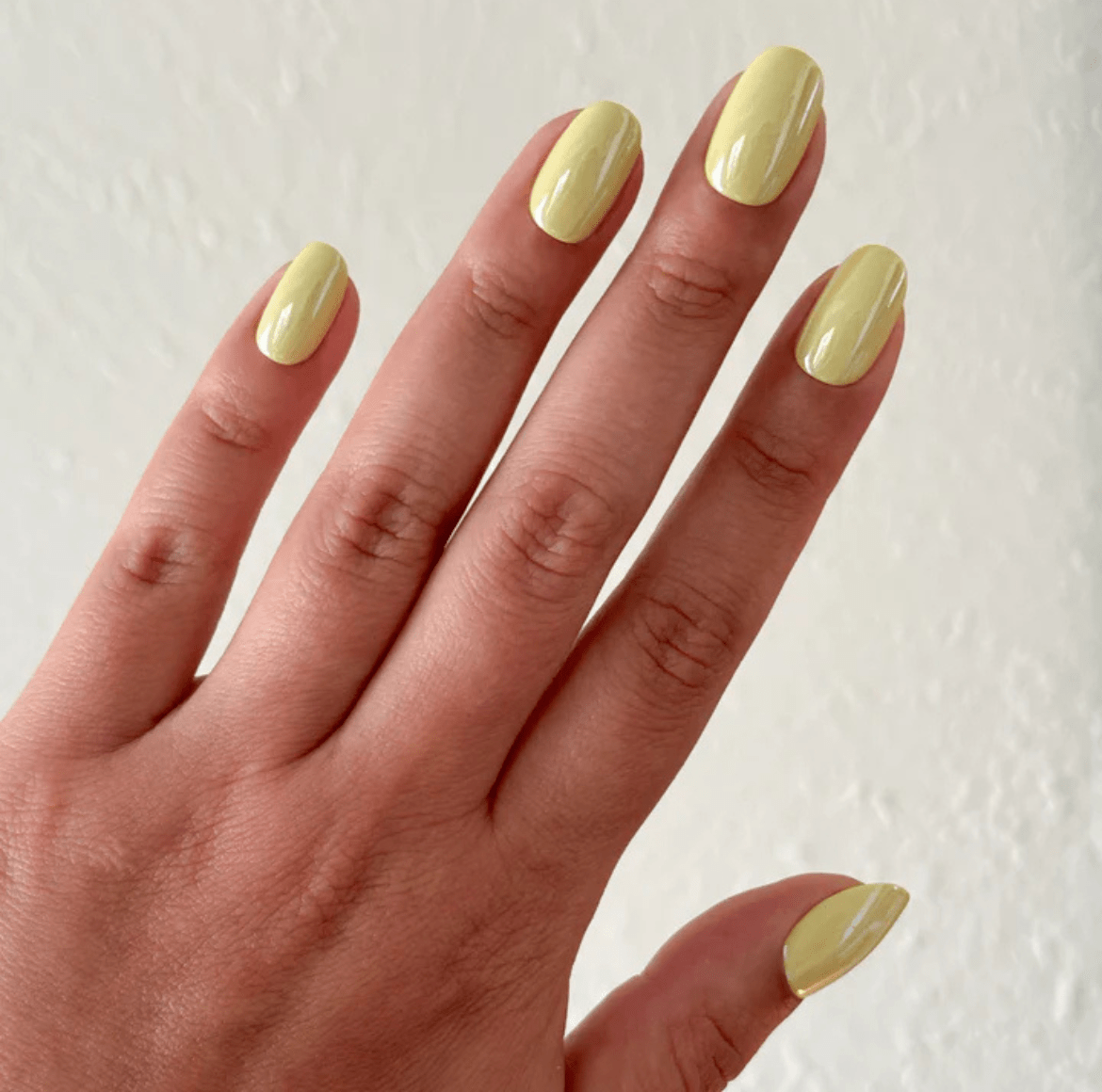 Close-up of a hand with pastel yellow chrome nails, featuring a glossy finish and natural skin texture against a neutral textured background