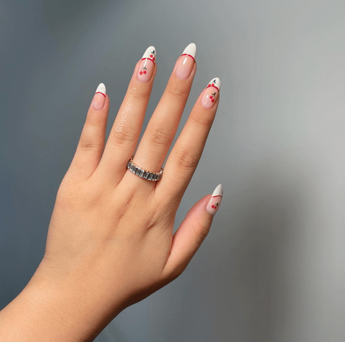 Almond-shaped nails with white tips, delicate red cherry designs, and a silver ring with rectangular stones on the index finger.