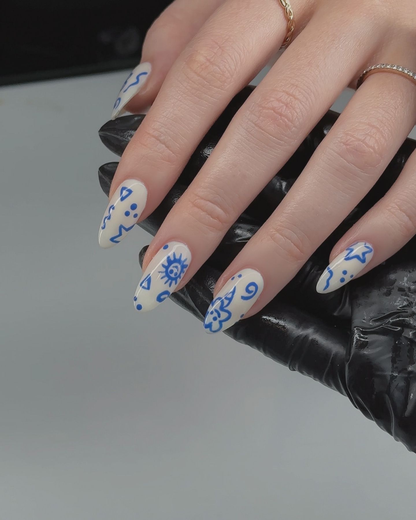 Almond-shaped nails painted creamy pale with cute blue doodle patterns, paired with delicate sparkly rings on the middle and ring fingers, set against a simple background.
