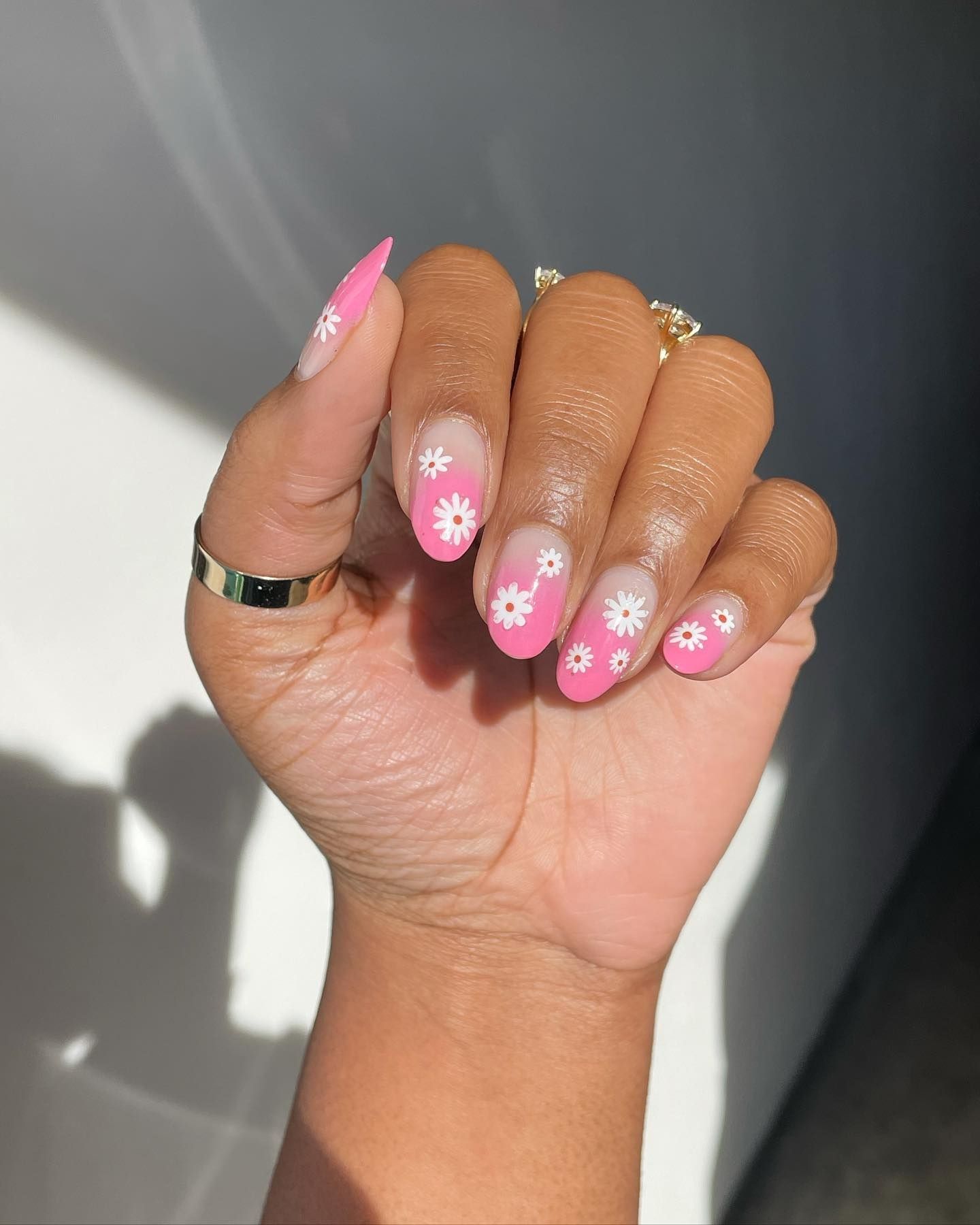 Almond-shaped pink gradient nails with hand-painted white daisies and gold rings on a sunlit hand