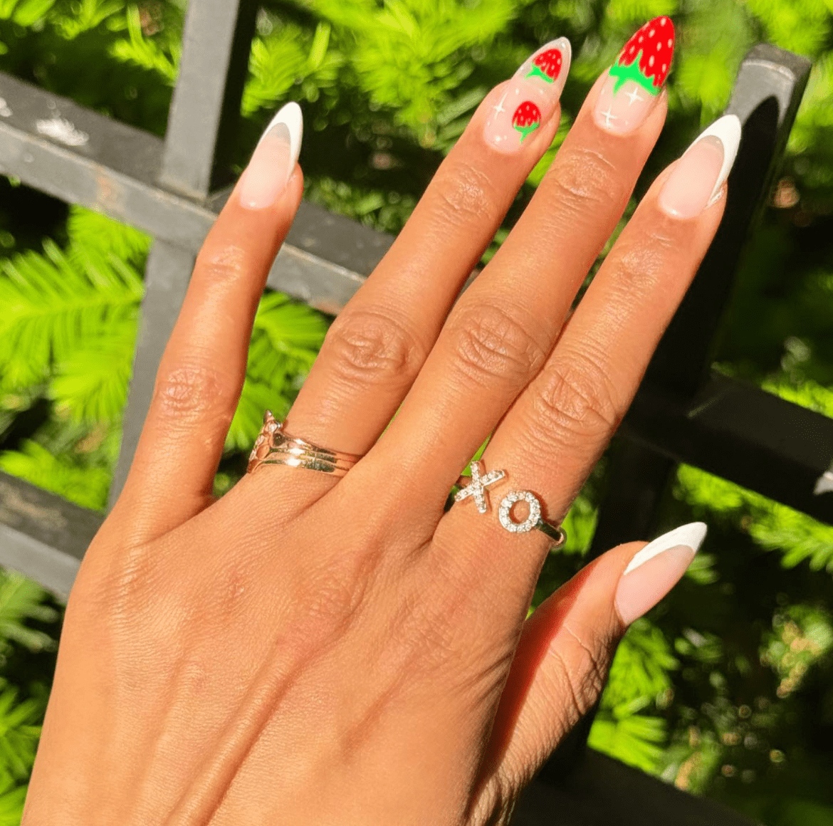 Almond-shaped French tip nails with soft pink bases and white tips, styled with XO and crown rings, photographed outdoors in summer sunlight